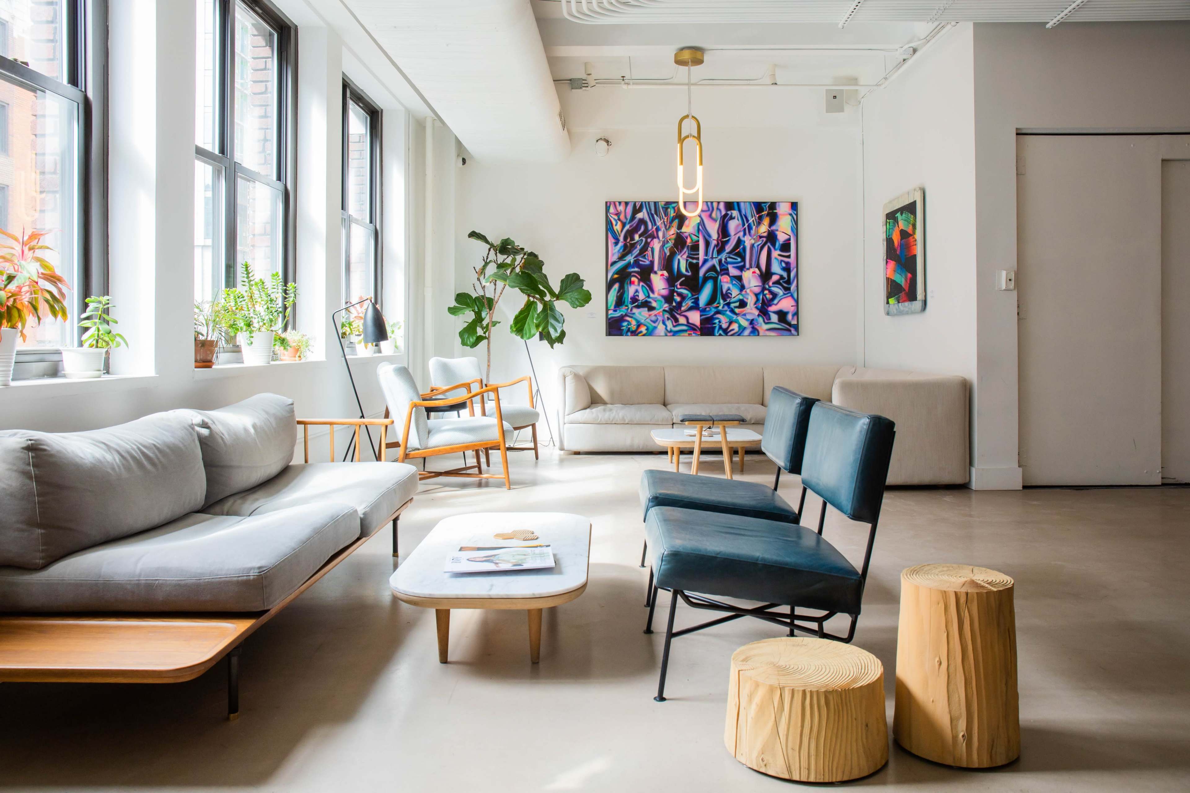 The image shows a modern, minimalist living room featuring various seating options, large windows, potted plants, and a colorful piece of artwork on the wall.