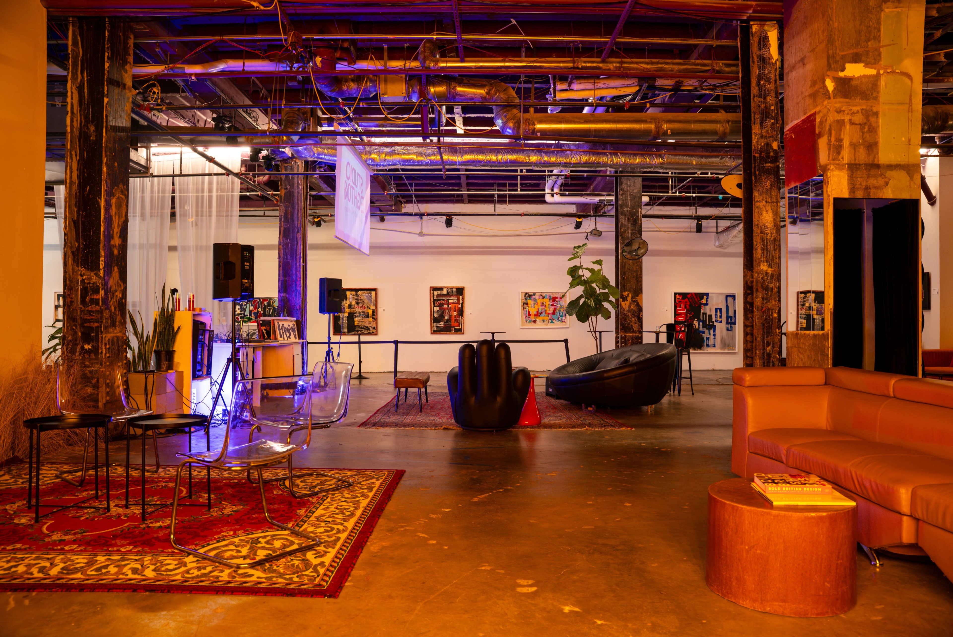 The image shows an interior space with a mix of modern furniture, artistic displays on the walls, and various lighting elements.