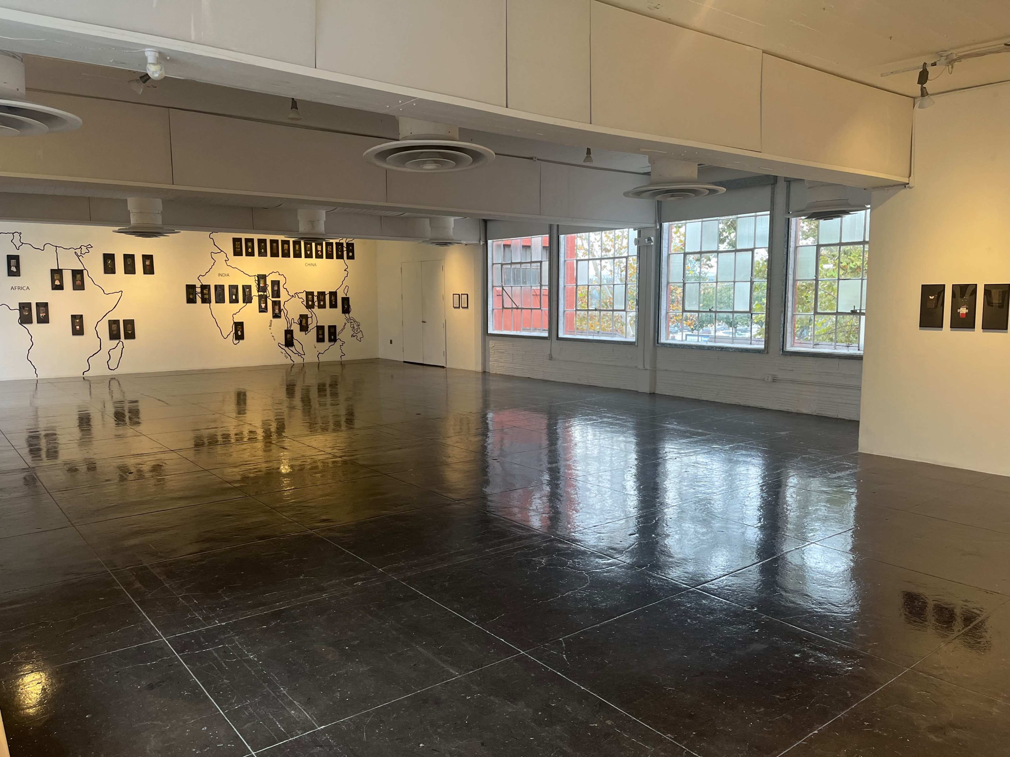 An empty art gallery with polished concrete floors, white walls, and a series of framed artworks hanging on one wall.