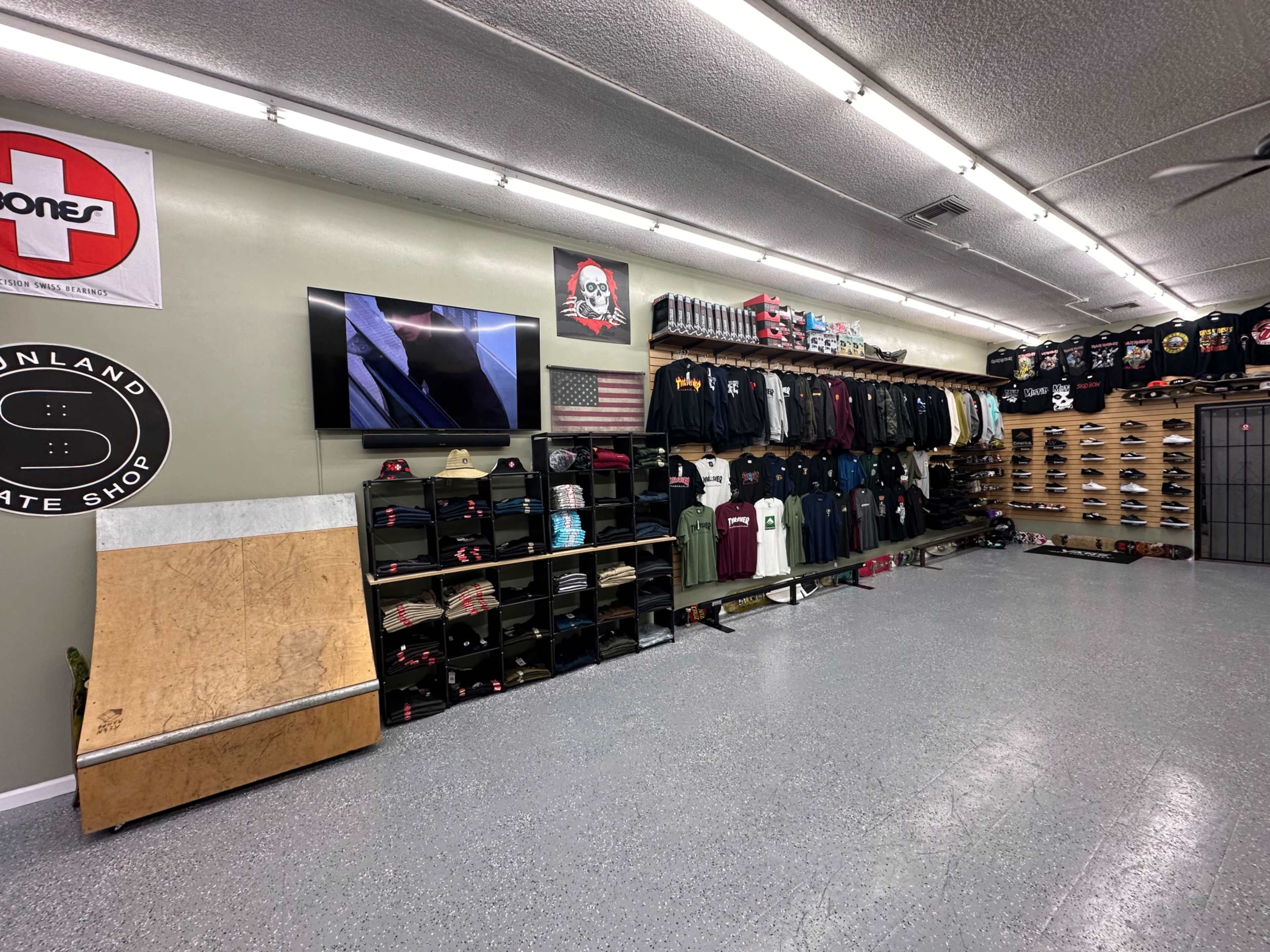 The interior of a skate shop with clothing and skateboards neatly arranged on shelves, a small ramp in the foreground, and a television mounted on the wall.
