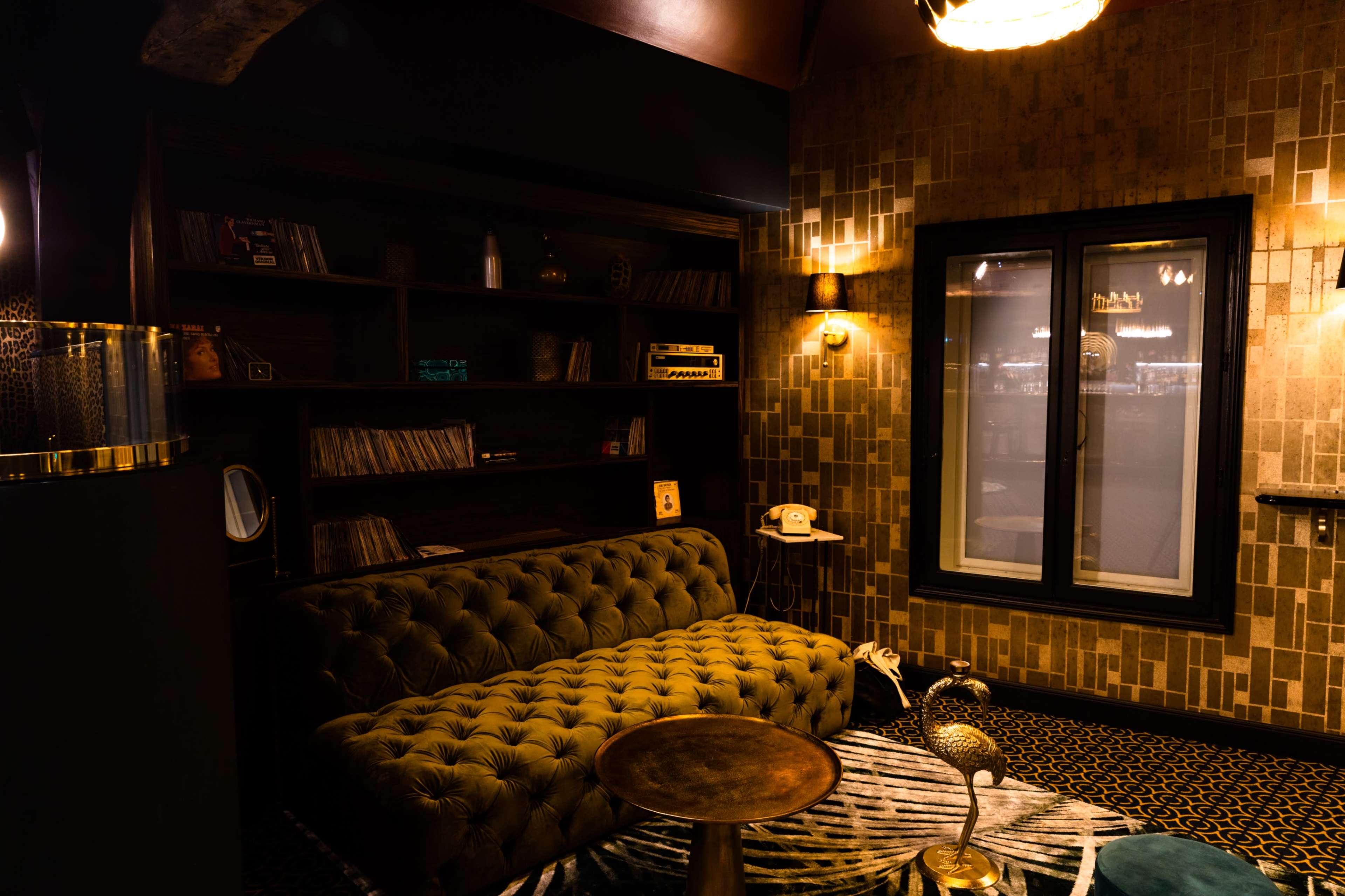 The image shows a cozy living space featuring a vintage sofa, a small metal table, and a wall-mounted bookshelf filled with records and decor items.