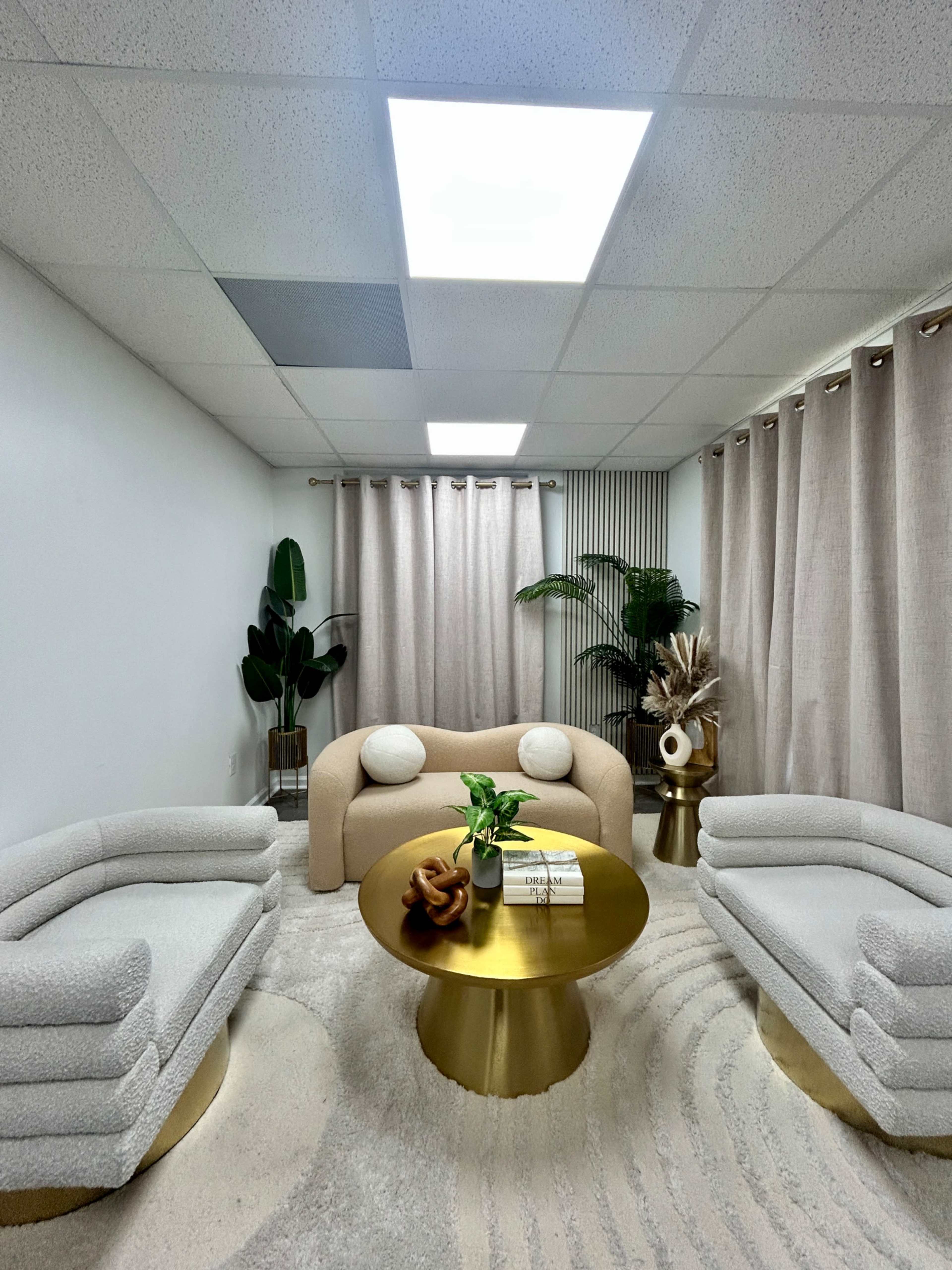 The image depicts a modern, minimalist lounge area with a beige sofa, two fluffy armchairs, a round gold coffee table, and decorative plants, all set against soft neutral curtains.