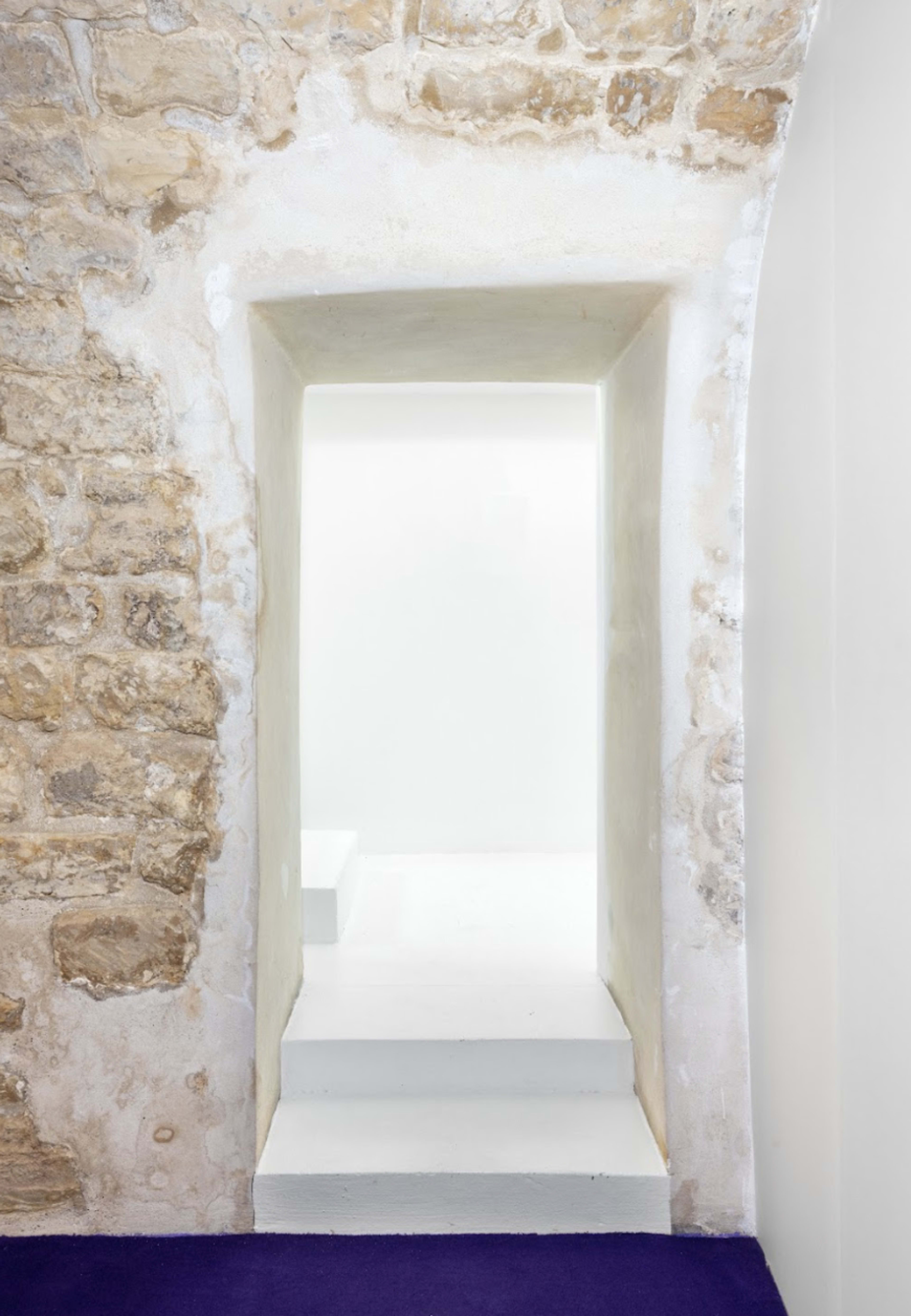 A simple archway leads from a stone wall into a brightly lit, minimalistic space with white steps.