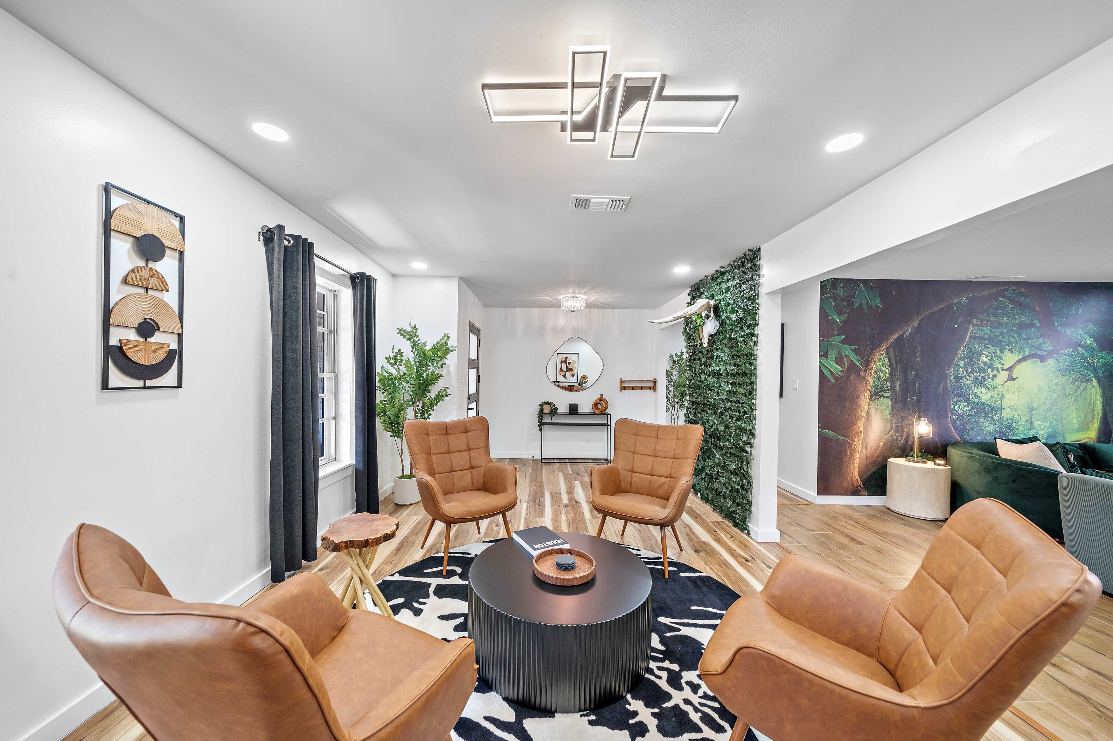 A modern living room featuring four tan chairs arranged around a black circular coffee table, with a green wall and a forest-themed mural in the background.
