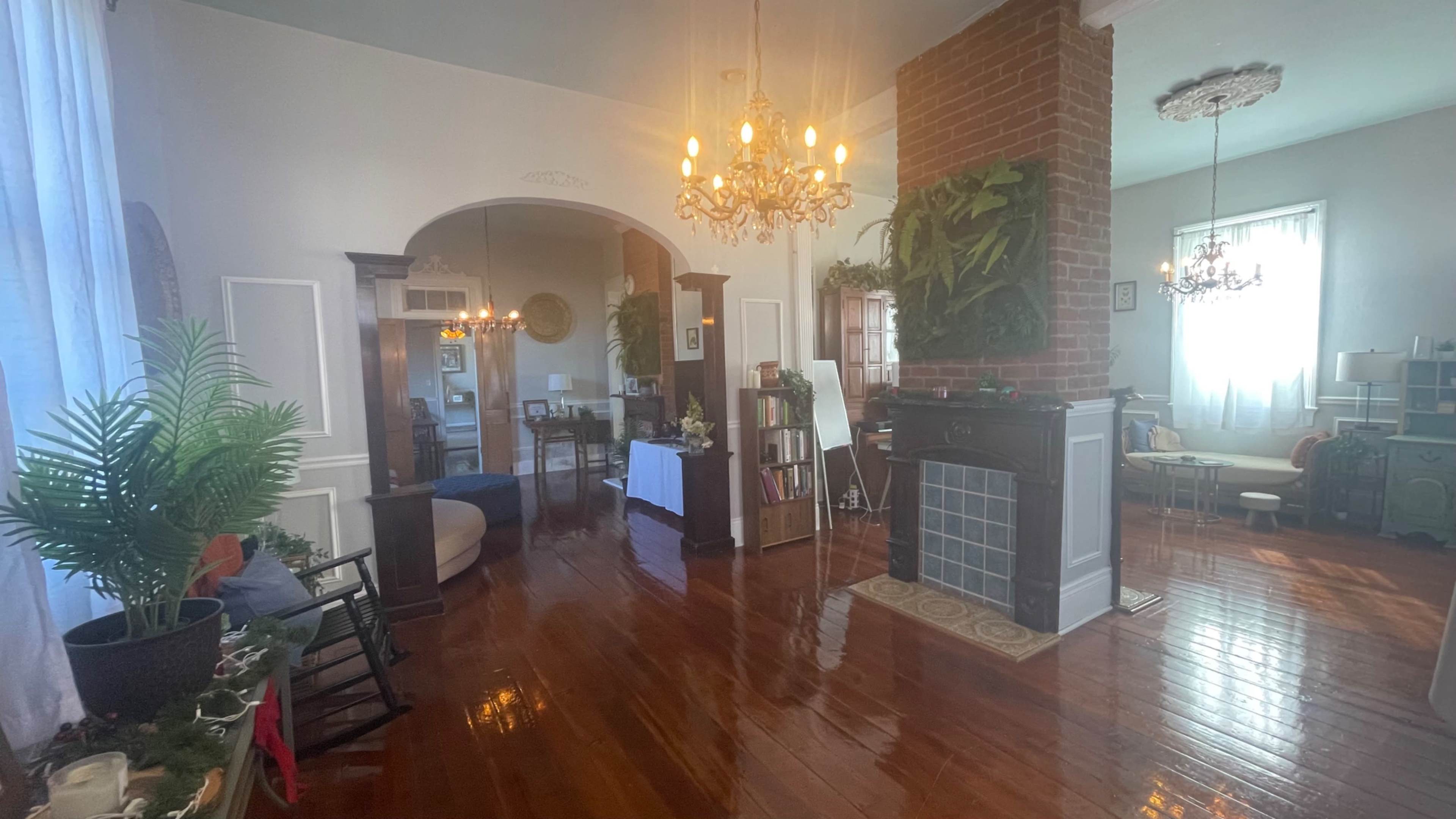 The image shows a spacious interior with polished wooden floors, featuring a living area with a fireplace, chandeliers, and various furnishings creating distinct seating and dining spaces.