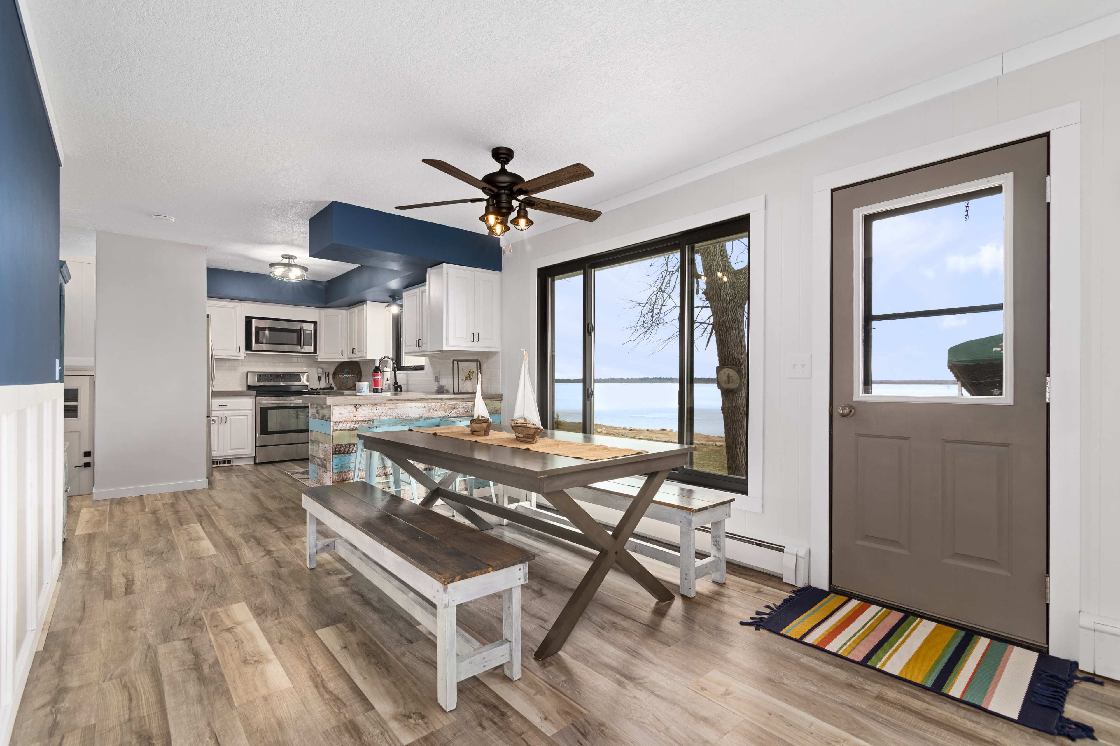 A light-filled kitchen and dining area with a large window overlooking a body of water, featuring a table with benches and a brown front door.