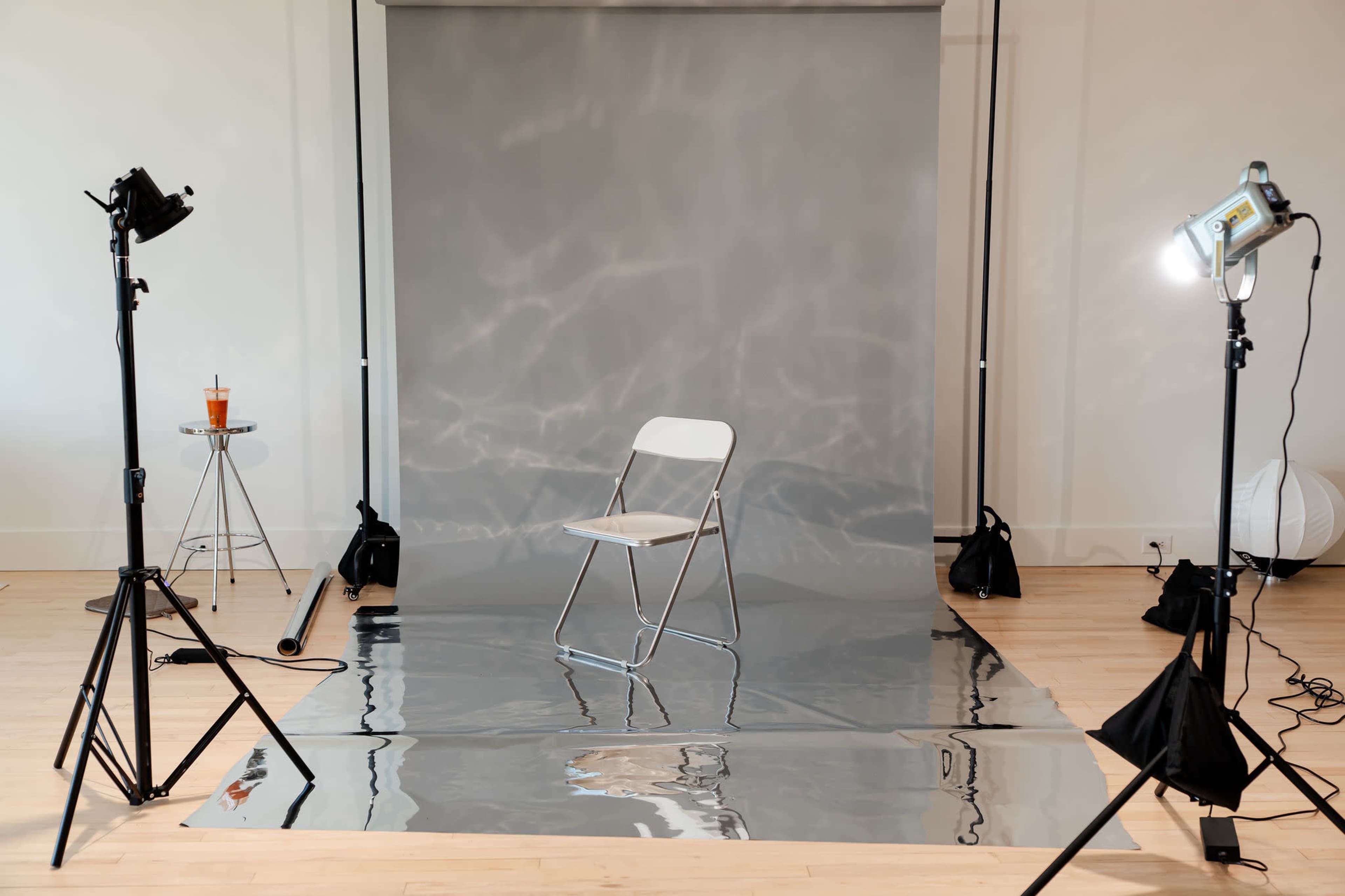The scene features a simple photography studio setup with a silver backdrop, a folding chair in the center, and lighting equipment on either side.