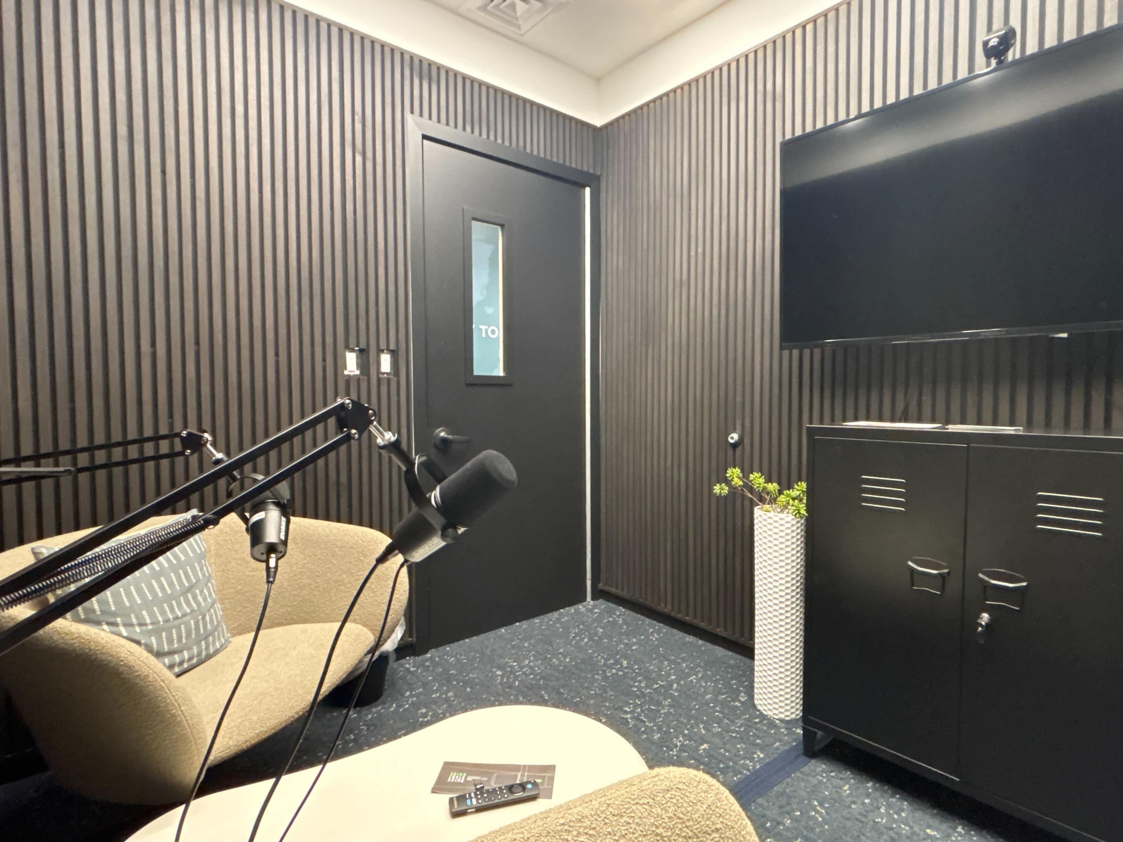 The image shows a small, modern meeting room with two chairs, a wall-mounted television, and a black cabinet against a textured wall.