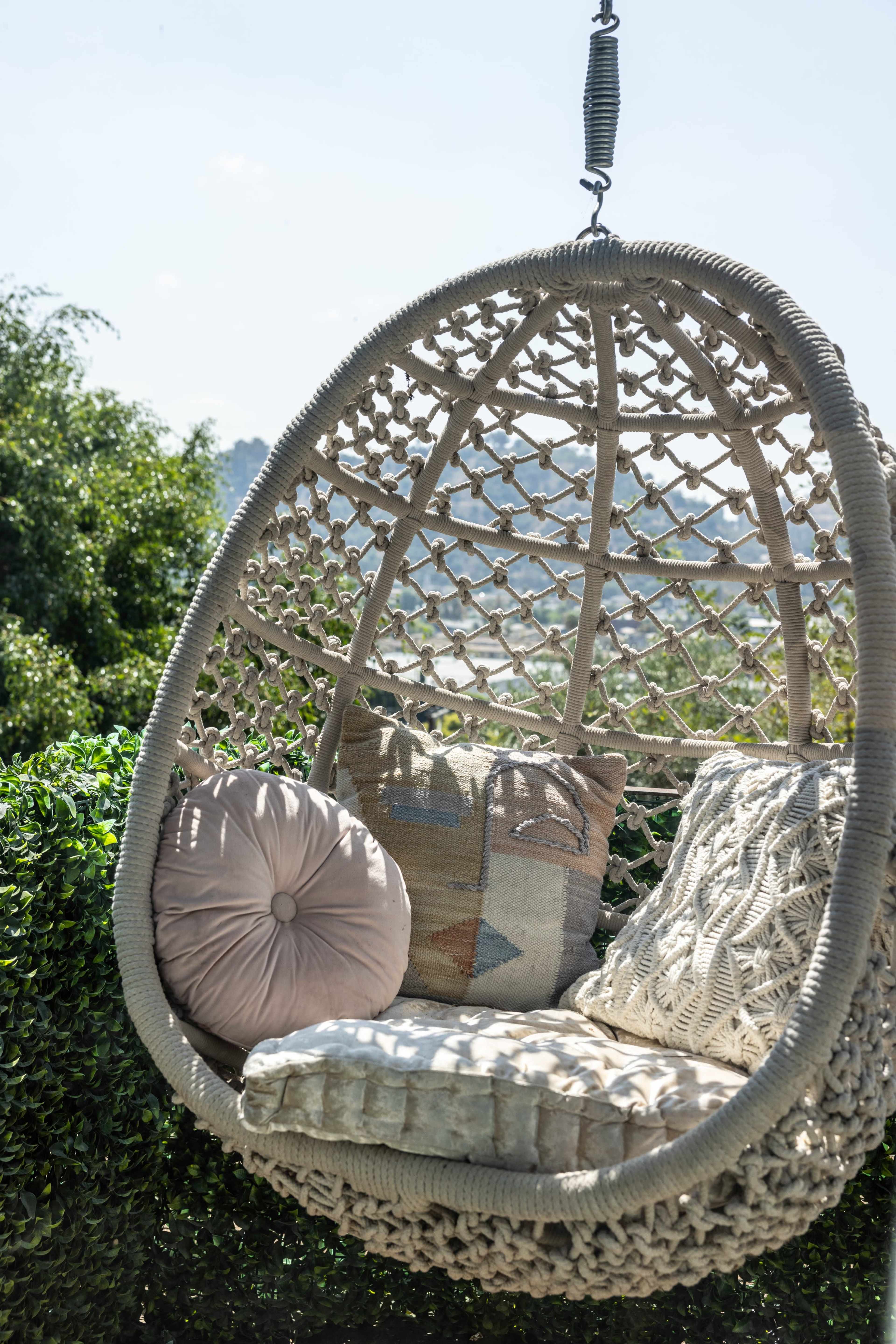 A hanging wicker chair with cushions is suspended among lush greenery, providing a cozy seating area outdoors.
