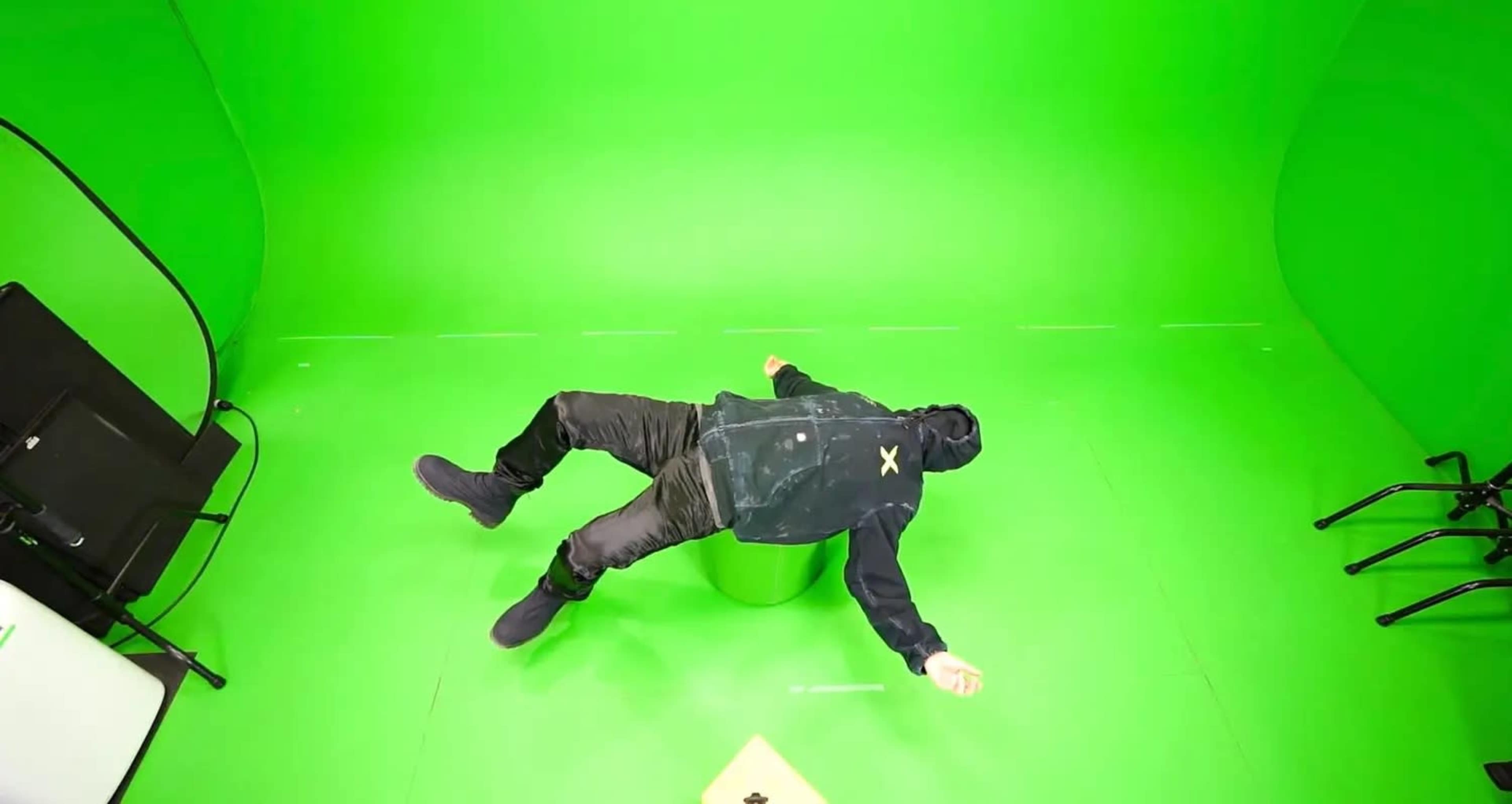 A person in dark clothing is lying horizontally over a reflective surface in a green-screen studio.