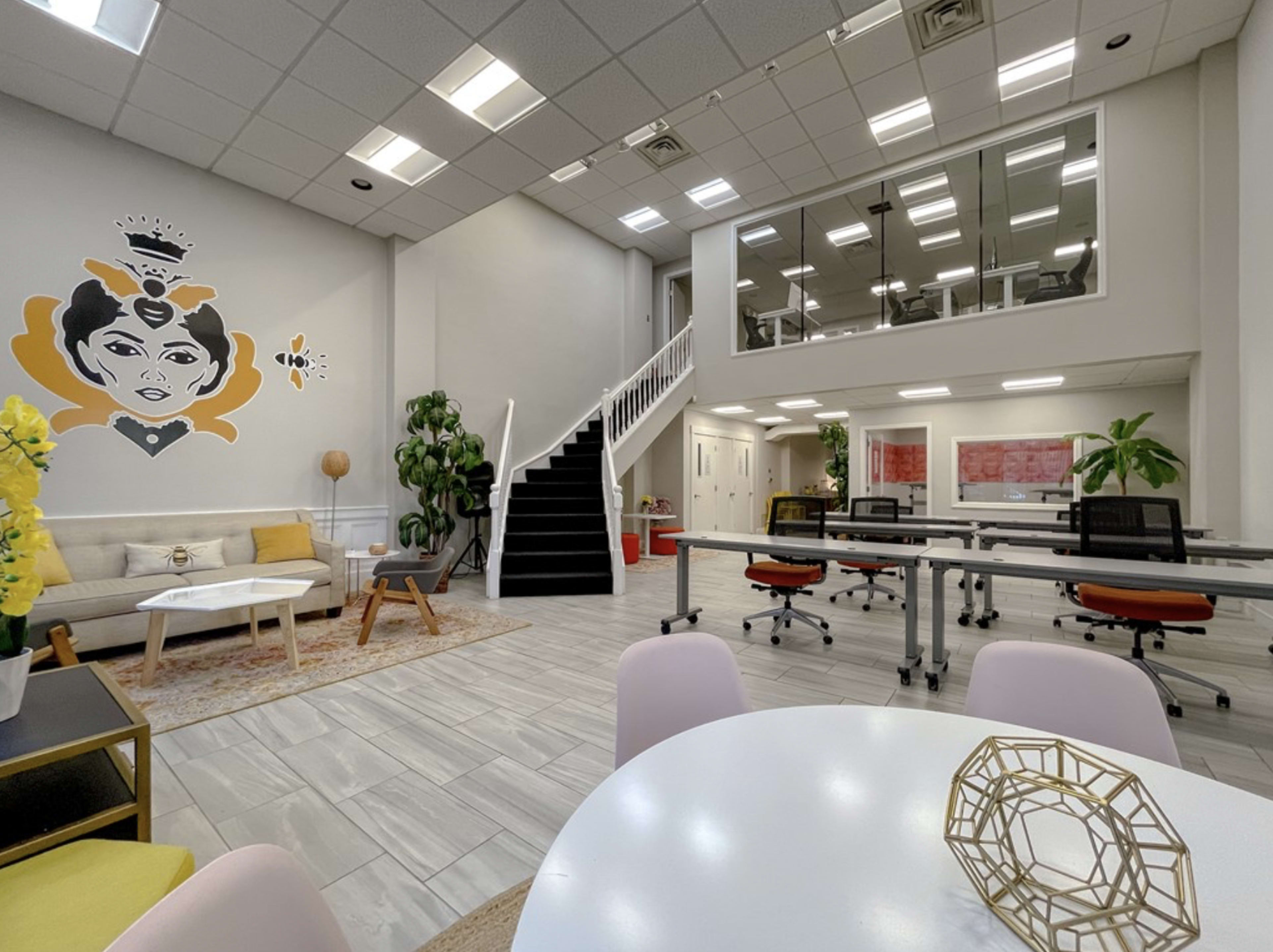 A modern office space features a seating area with a decorative mural, large windows, and an upper-level office with a black staircase.