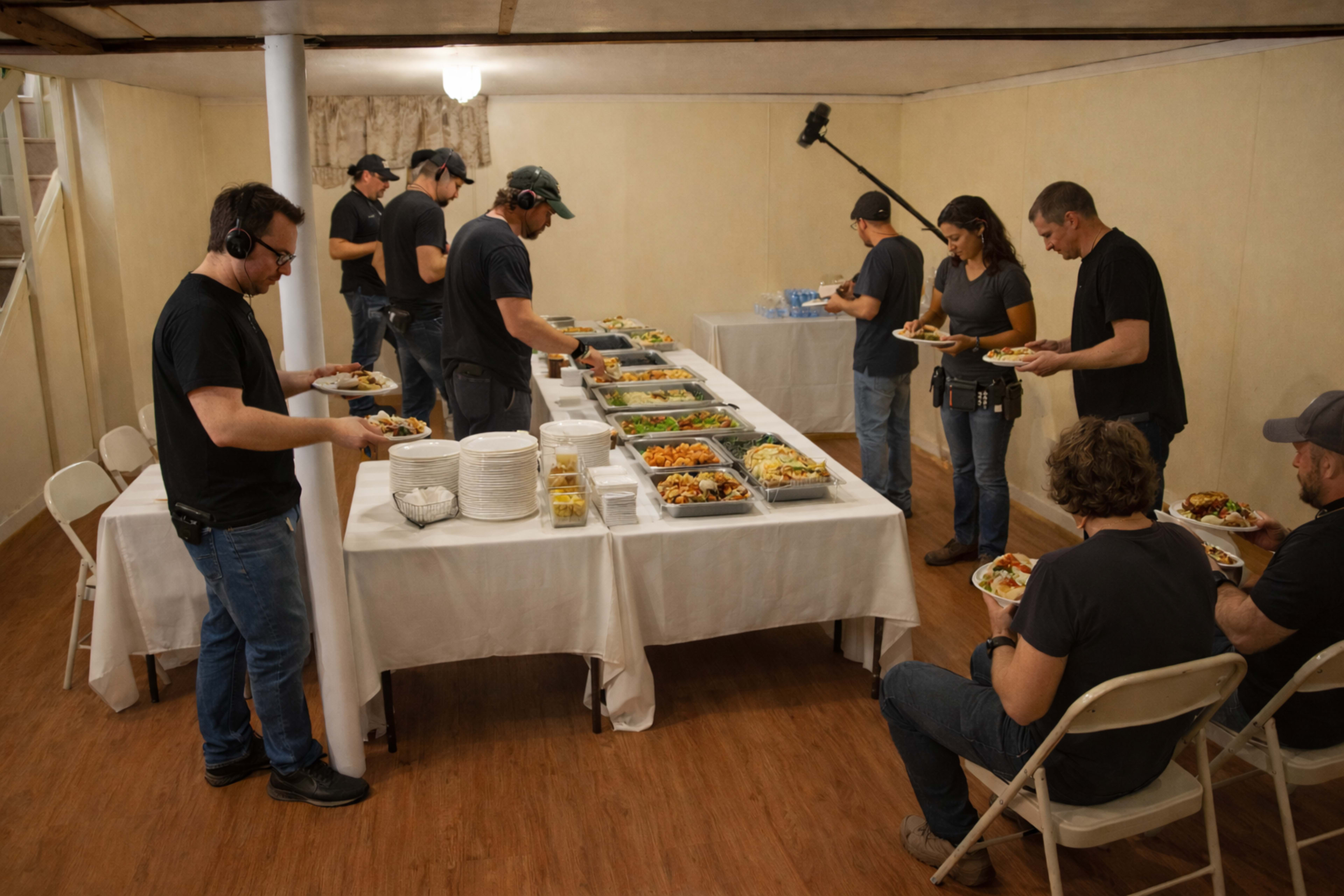A group of people is serving and enjoying a buffet meal in a basement setting, with tables covered in food and plates.