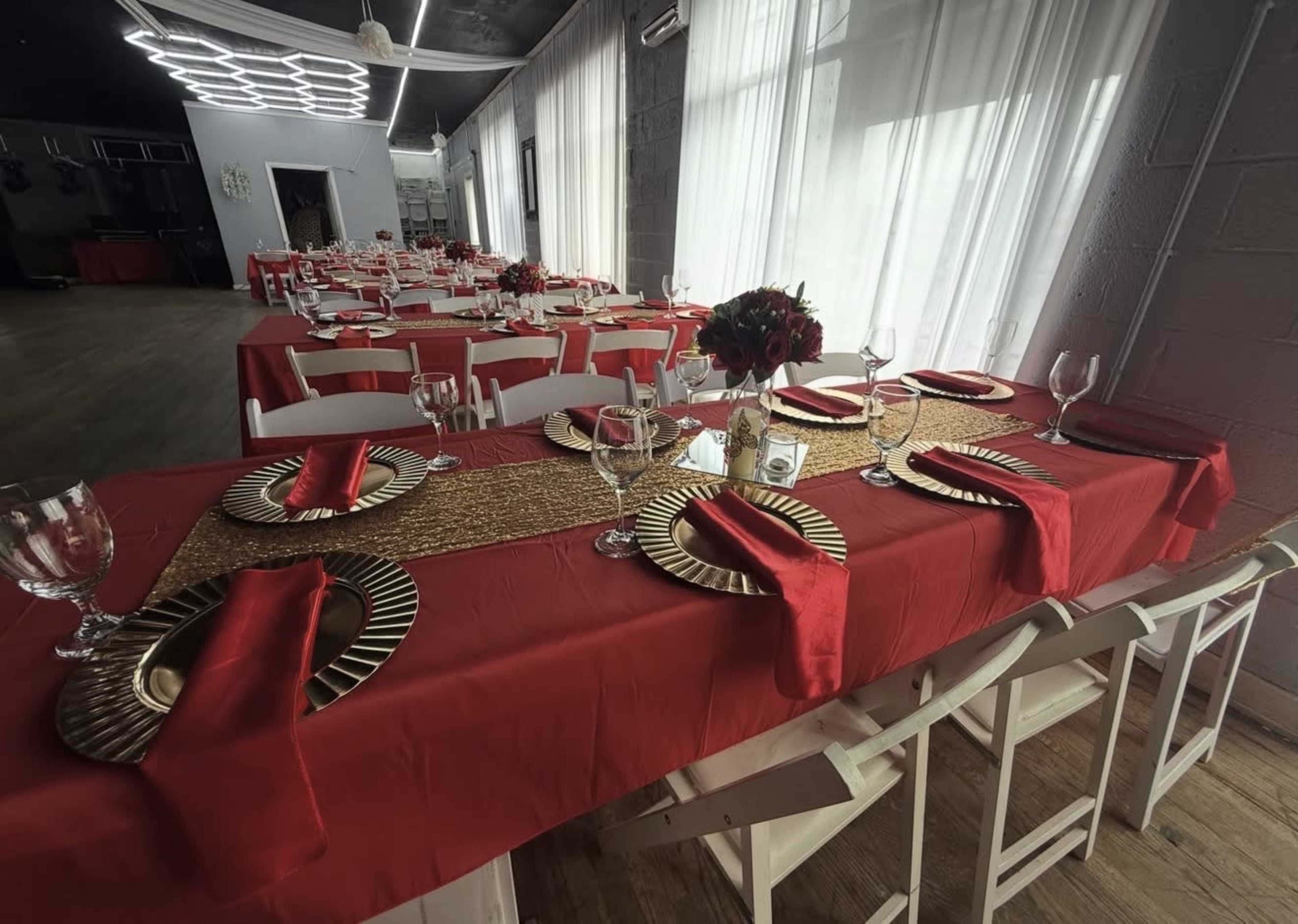 The image shows a dining area set for an event with tables covered in red tablecloths and decorated with gold plates and wine glasses.