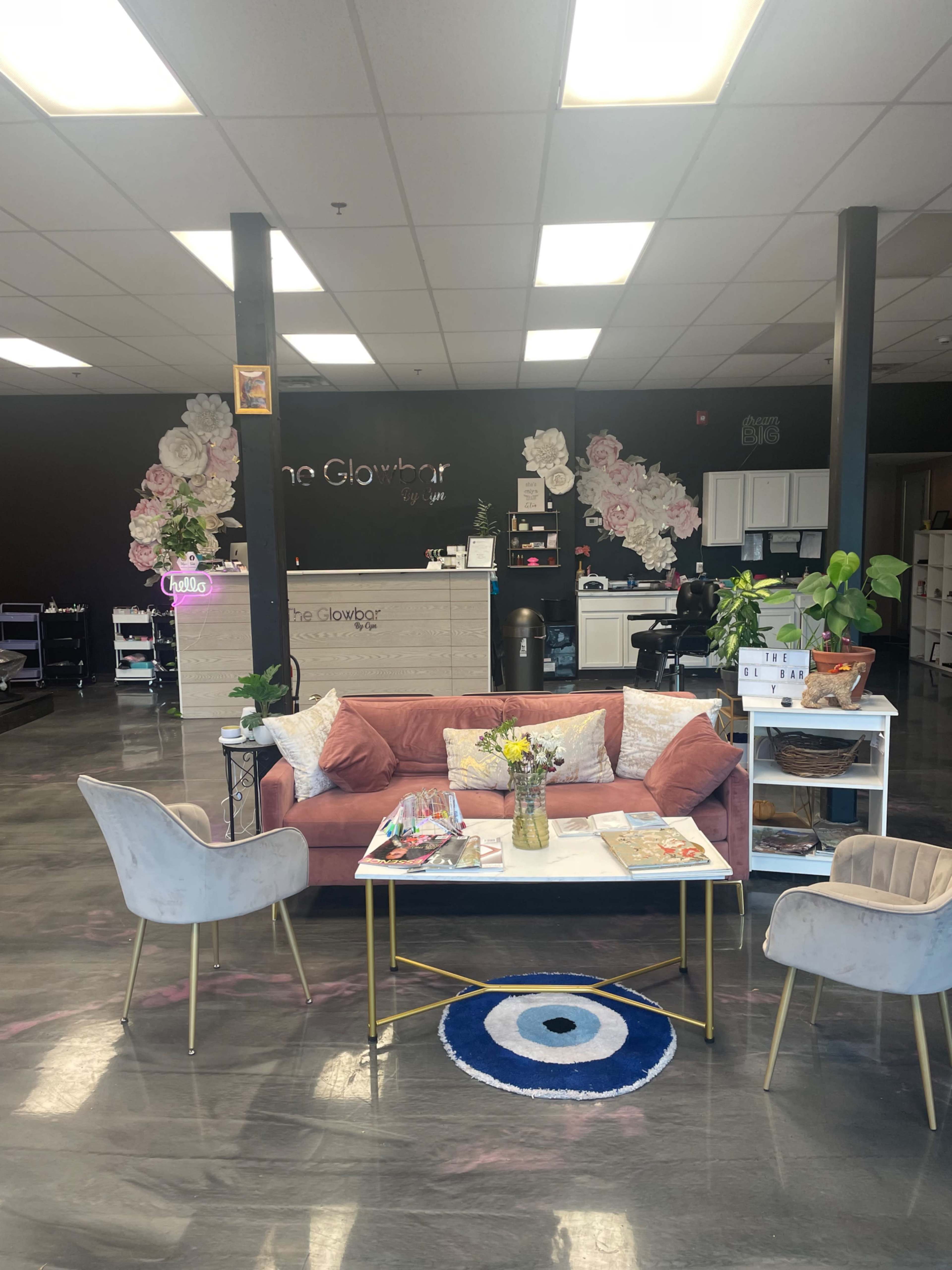 A modern beauty salon reception area with a pink couch, a coffee table, decorative flowers on the wall, and stylish chairs.