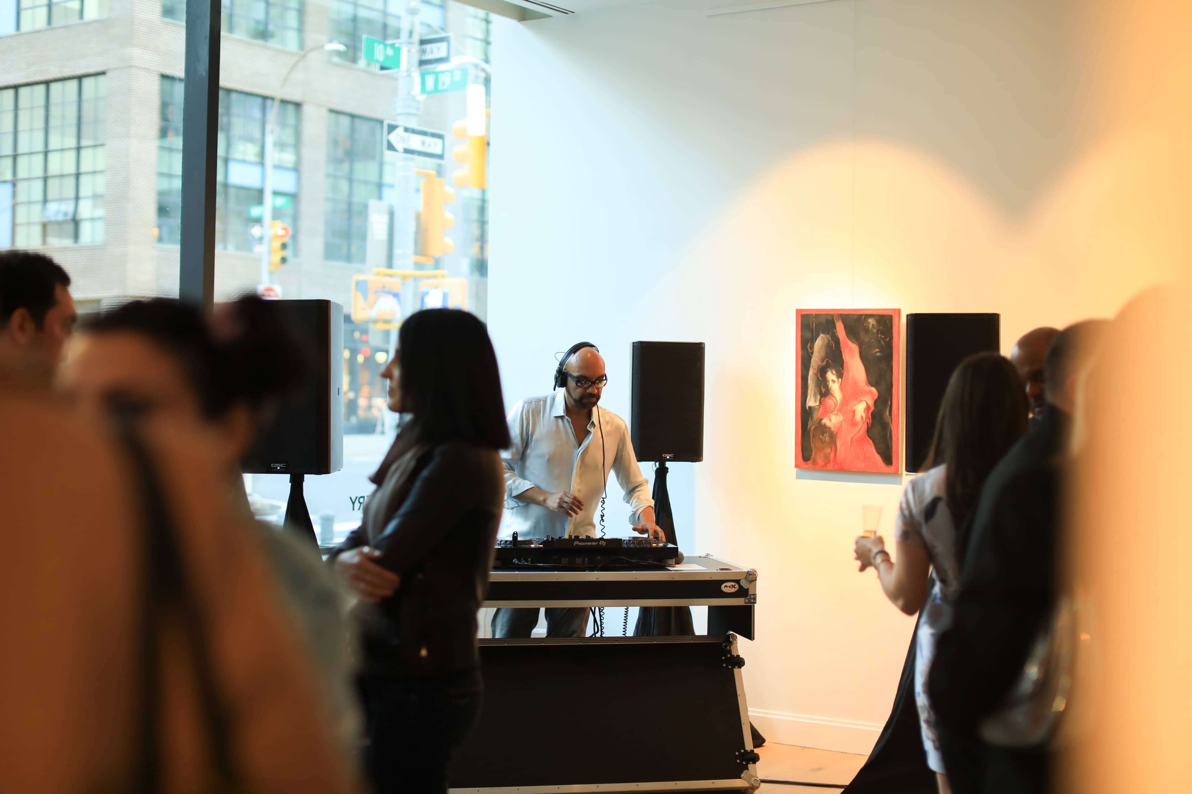 A DJ stands behind a turntable setup in an art gallery while attendees mingle in the foreground.