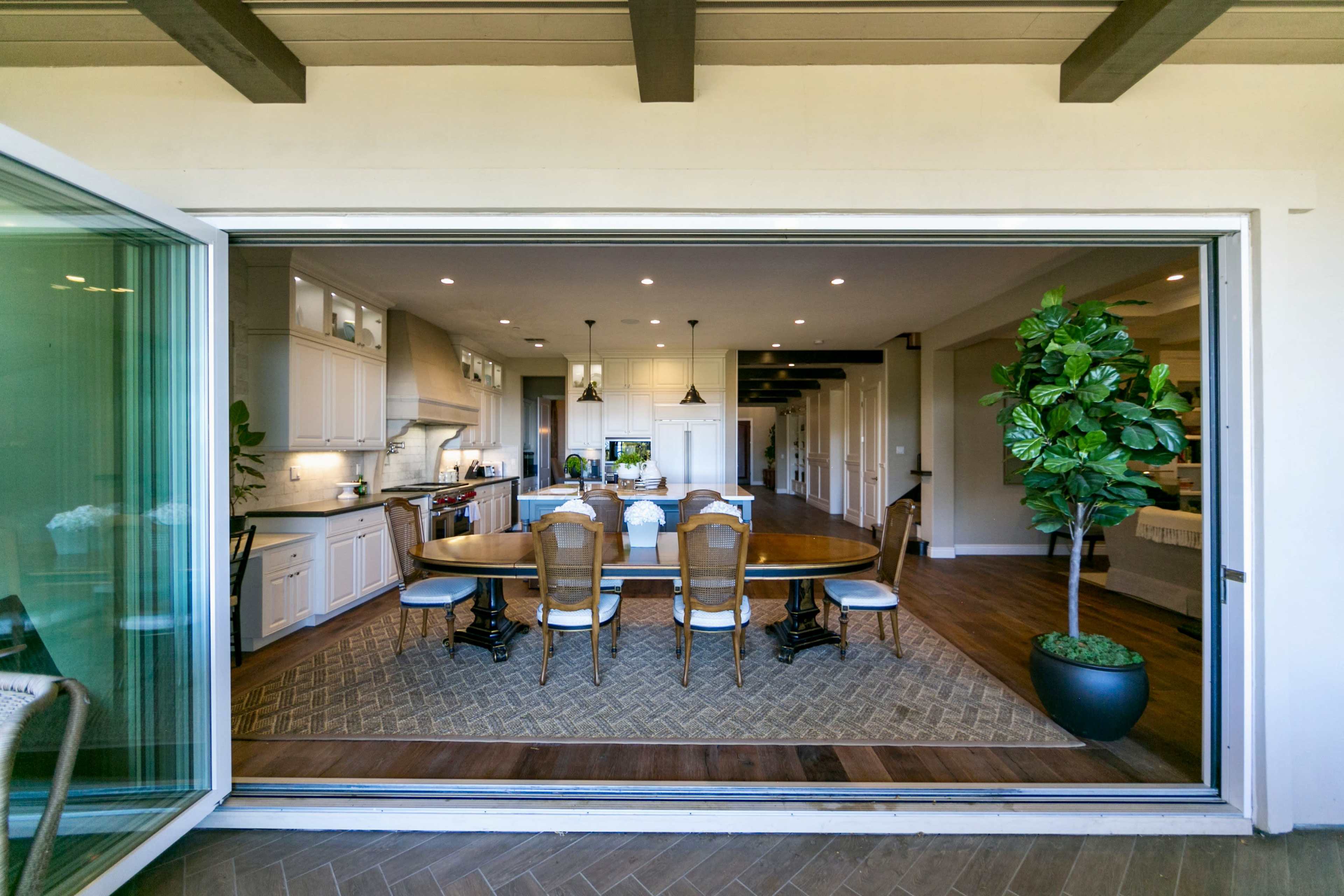 A spacious kitchen and dining area with a large wooden table, stylish chairs, and a potted plant, visible through large sliding glass doors.