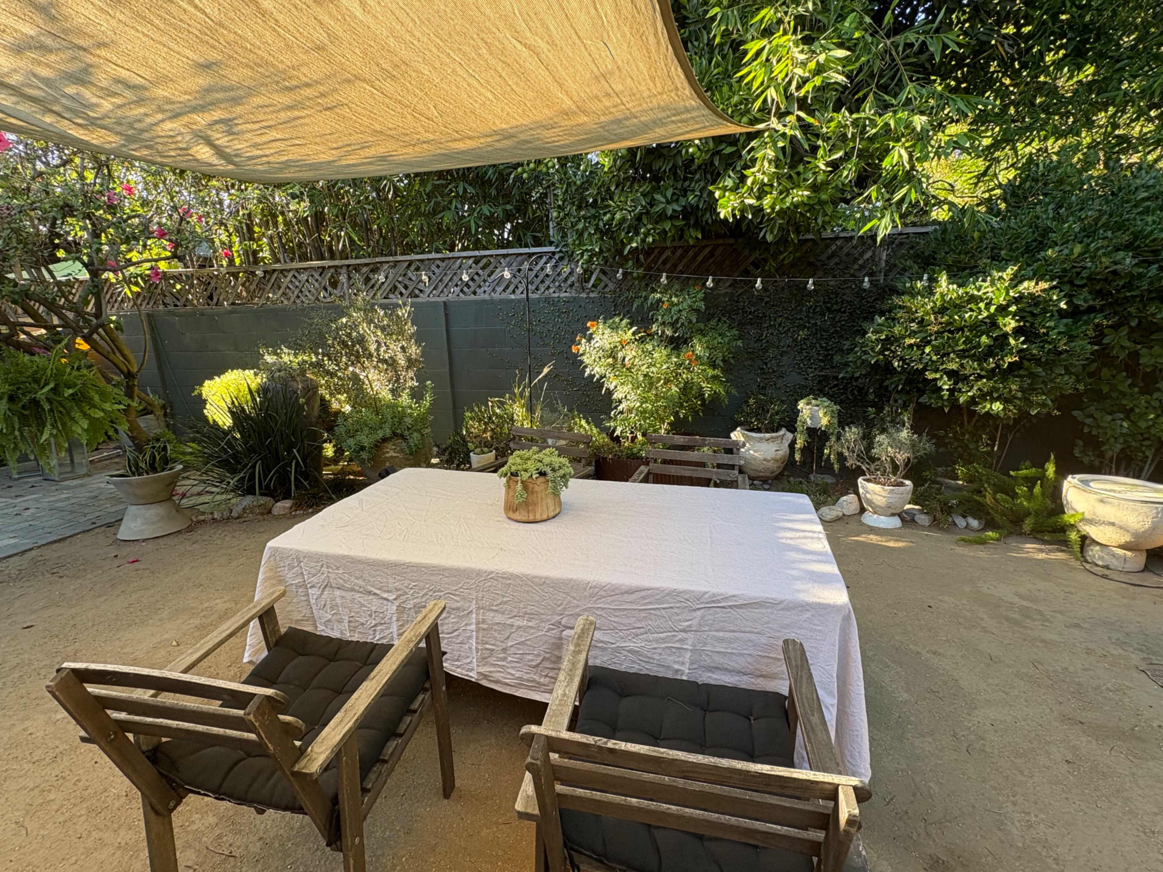 A shaded outdoor seating area features a table covered with a white tablecloth and two chairs, surrounded by various plants and greenery.