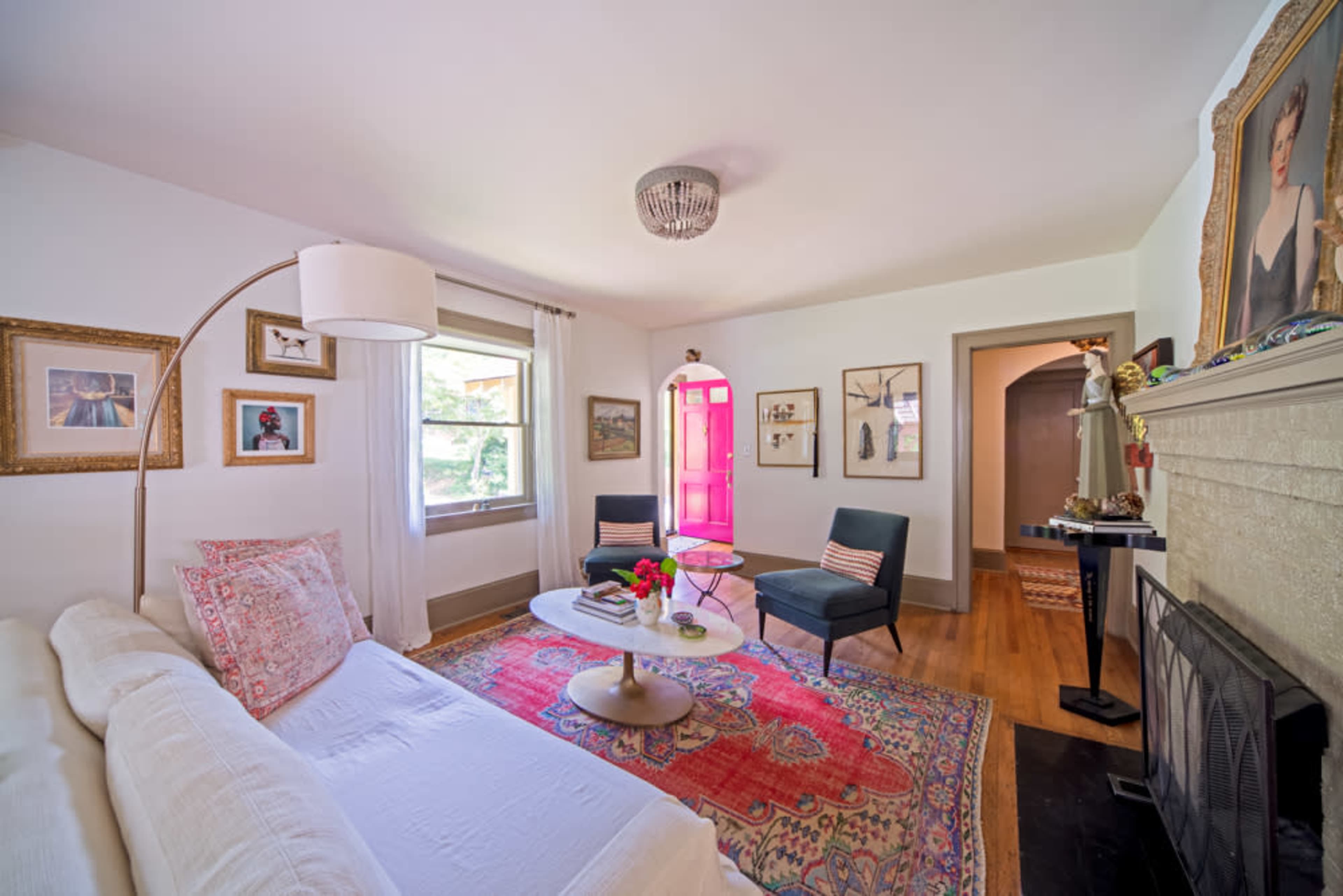 A bright and welcoming living room features a white sofa, two black chairs, a round coffee table, and colorful artwork on the walls, with a door painted bright pink.