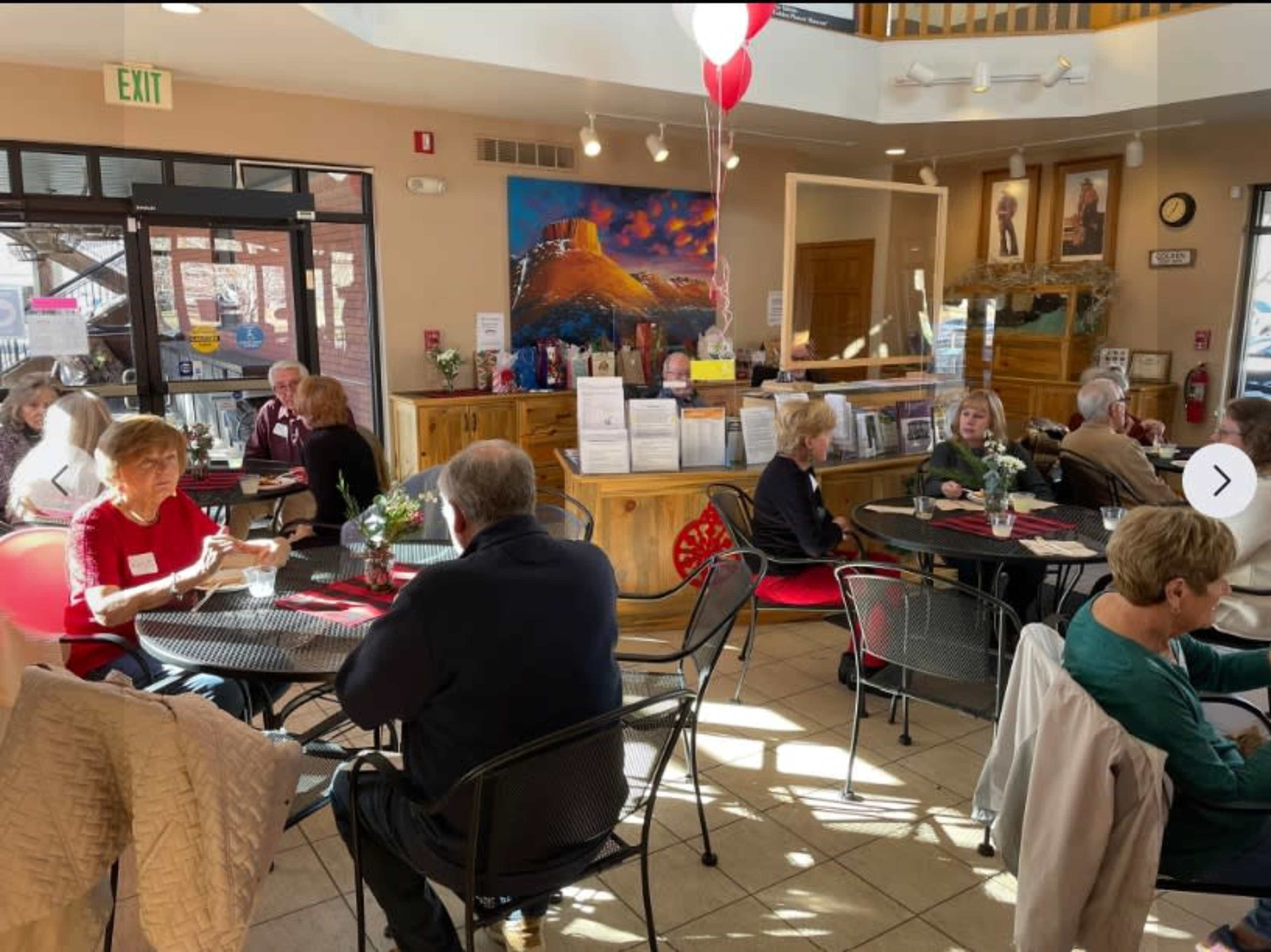 The image shows a busy café with several tables occupied by people engaging in conversation, surrounded by various decorations and a prominently displayed painting on the wall.