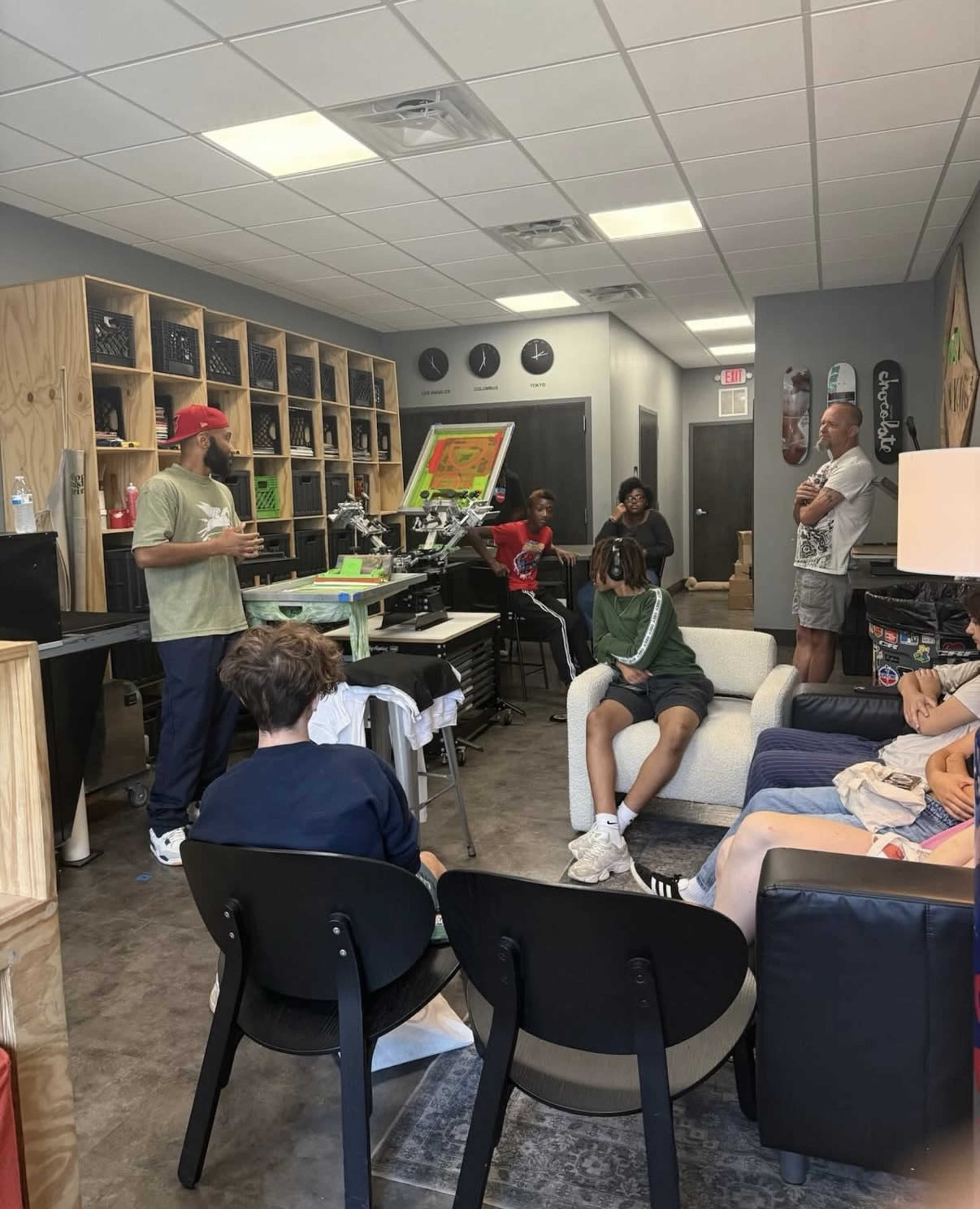 A group of individuals, including children and adults, are gathered in a room with screens and printing equipment while someone explains a process at the front.