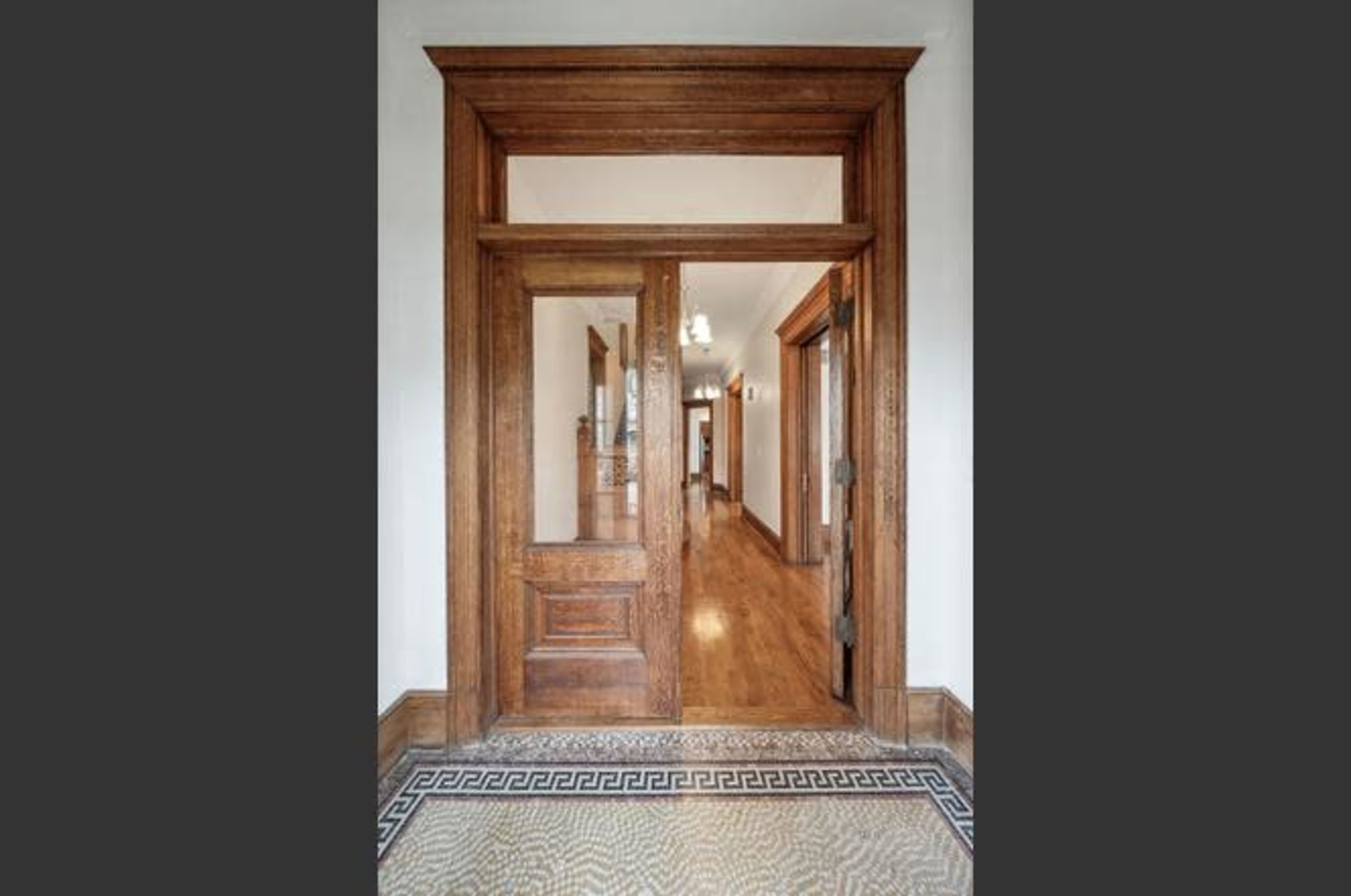The image shows a wooden entrance door leading into a corridor with hardwood floors and decorative tile.