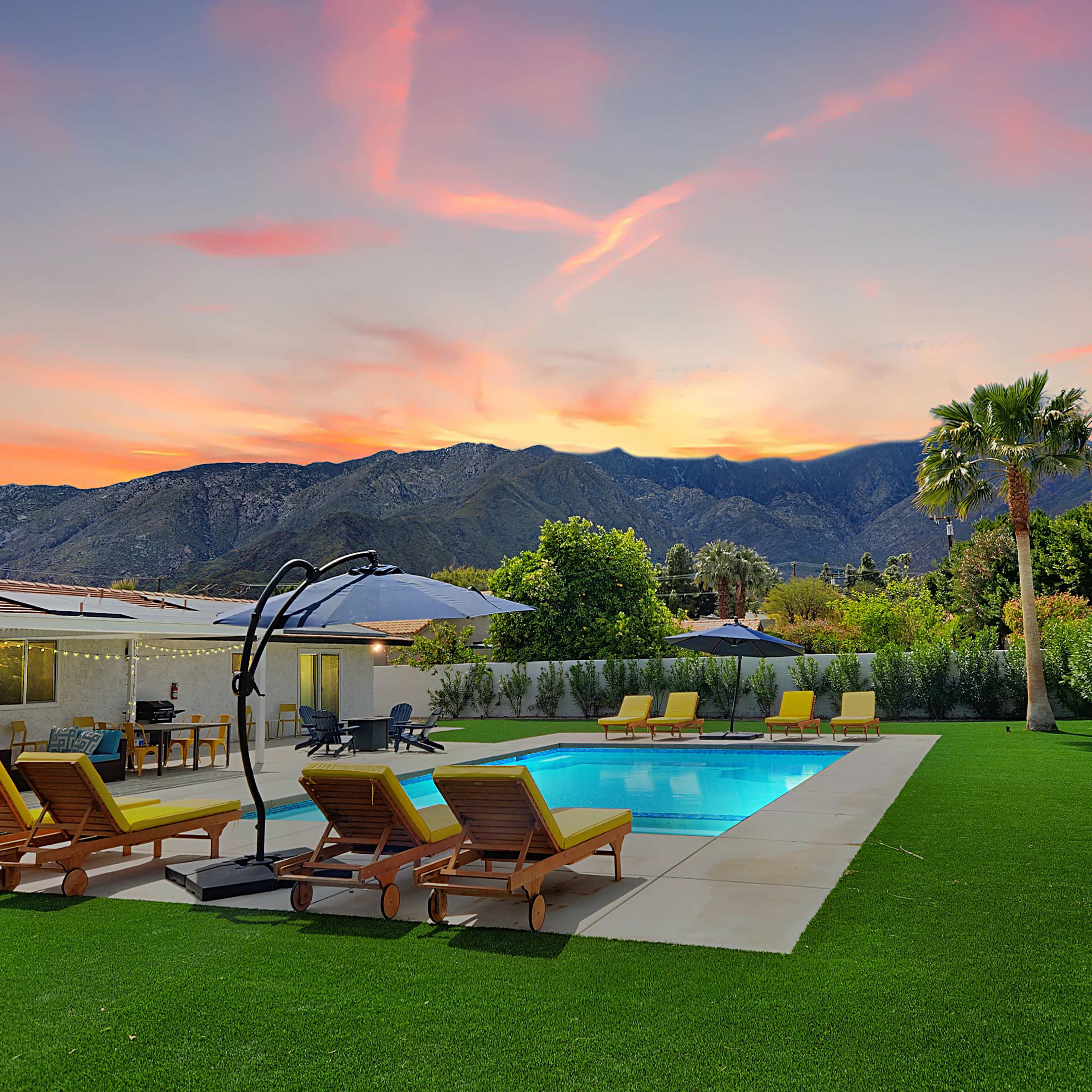 The image shows a swimming pool surrounded by lounge chairs and umbrellas, with mountains in the background under a colorful sunset.
