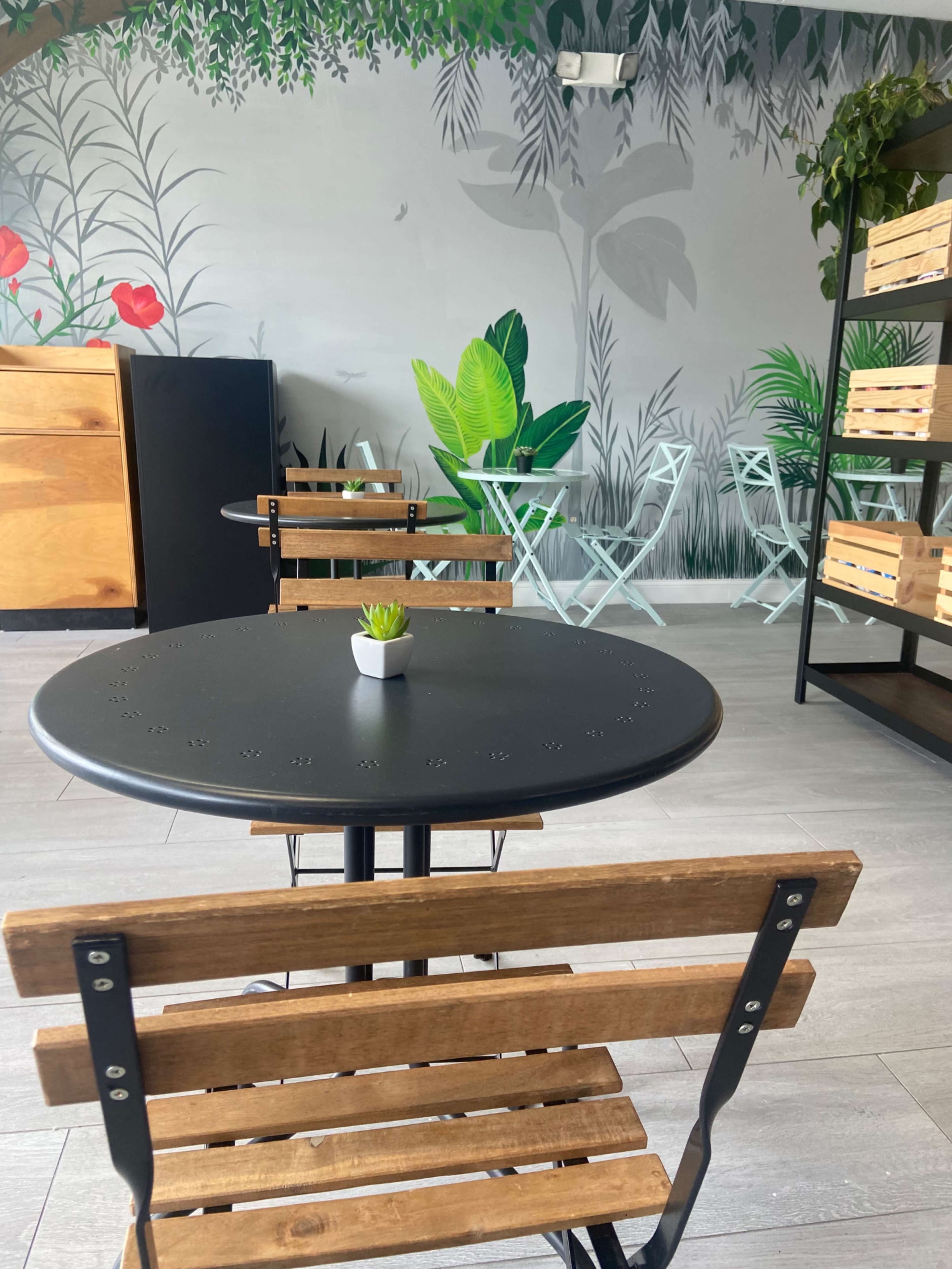 A small black round table with wooden chairs is set in a café space featuring a mural of greenery and plants on the wall.