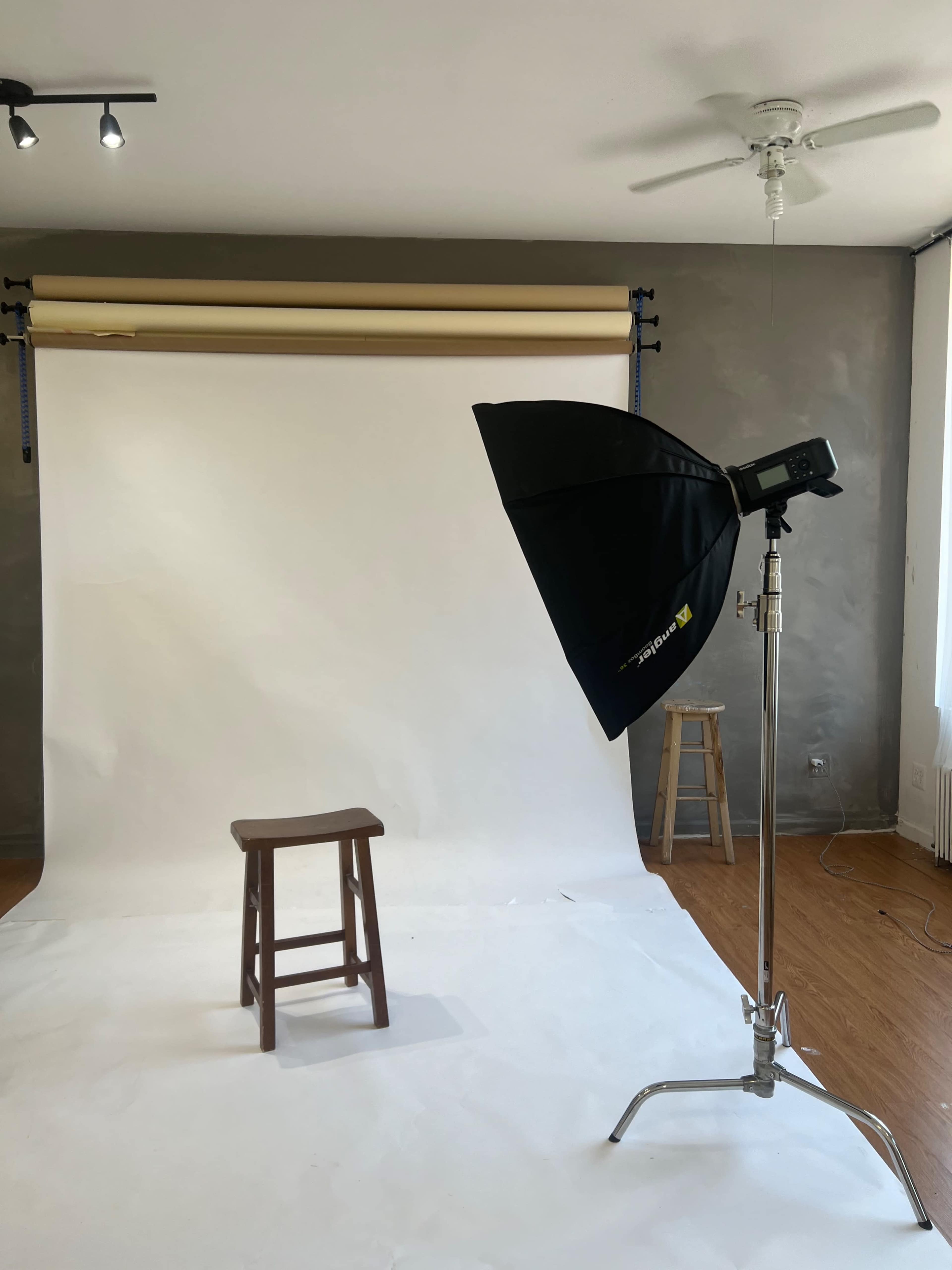 The image shows a photography studio setup with a wooden stool in front of a white backdrop and a softbox lighting on a stand.