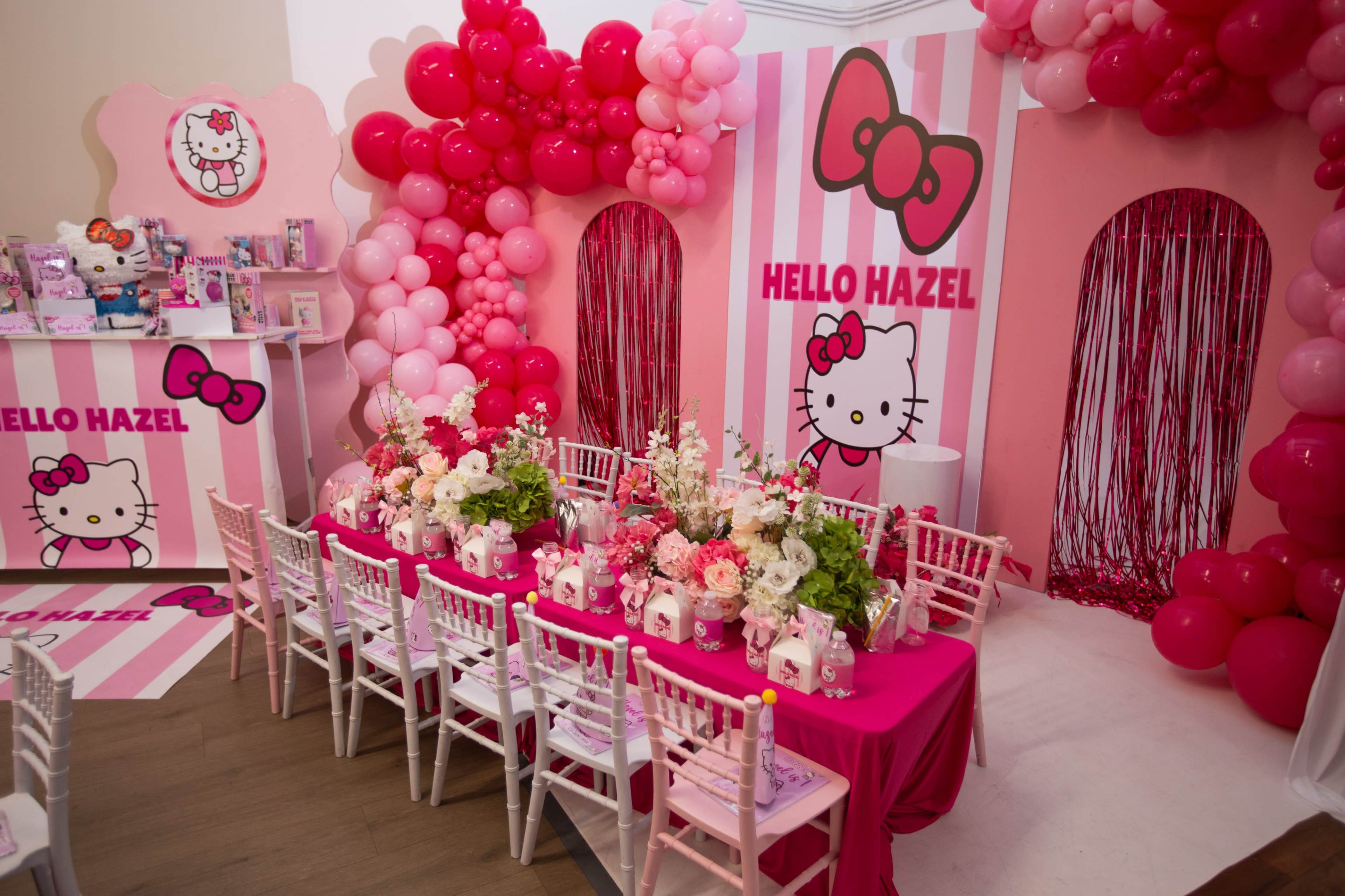 The image shows a themed party setting decorated in pink with Hello Kitty motifs, featuring a table adorned with flowers and party favors.