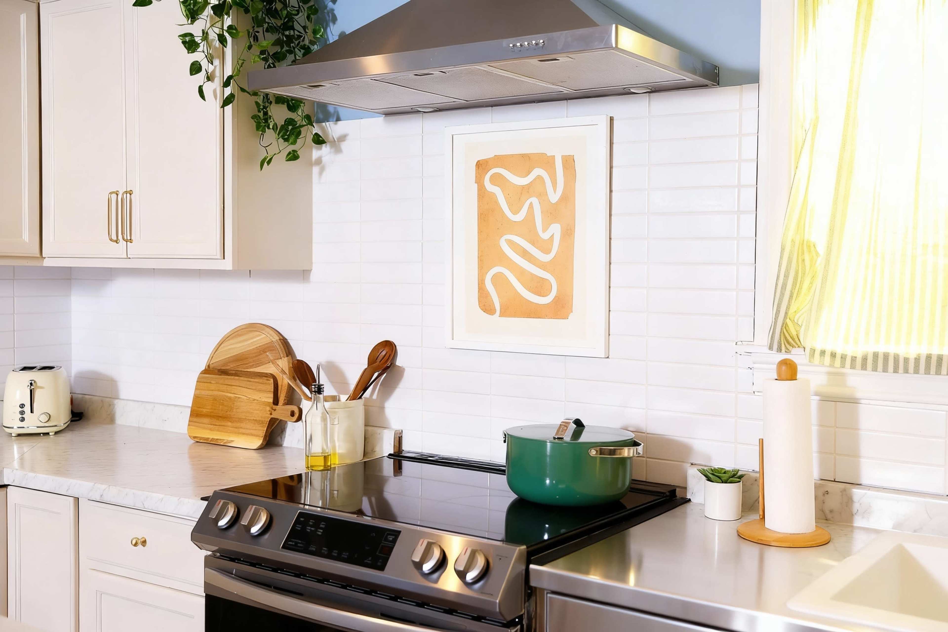 The image shows a modern kitchen with a stove, overhead range hood, wooden cutting board, and a decorative artwork on the wall.