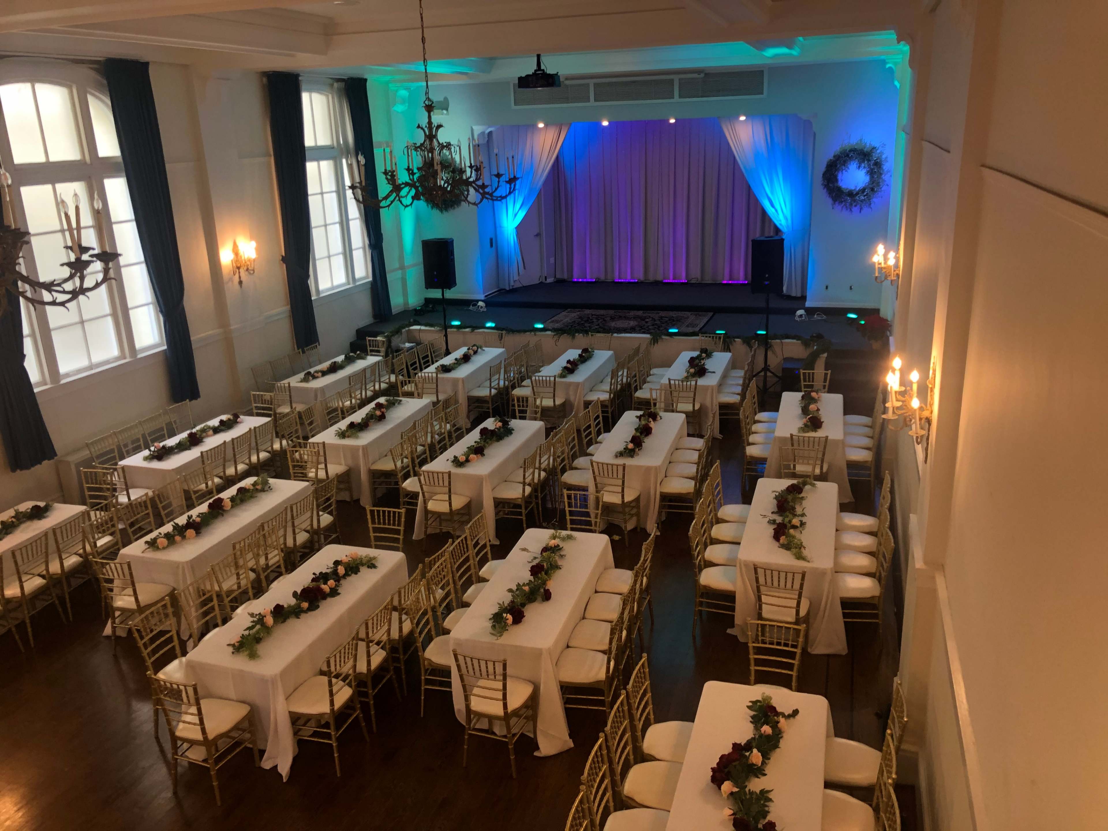 The room features rows of tables with white tablecloths and floral centerpieces, set up for an event underneath chandeliers and colored lights.