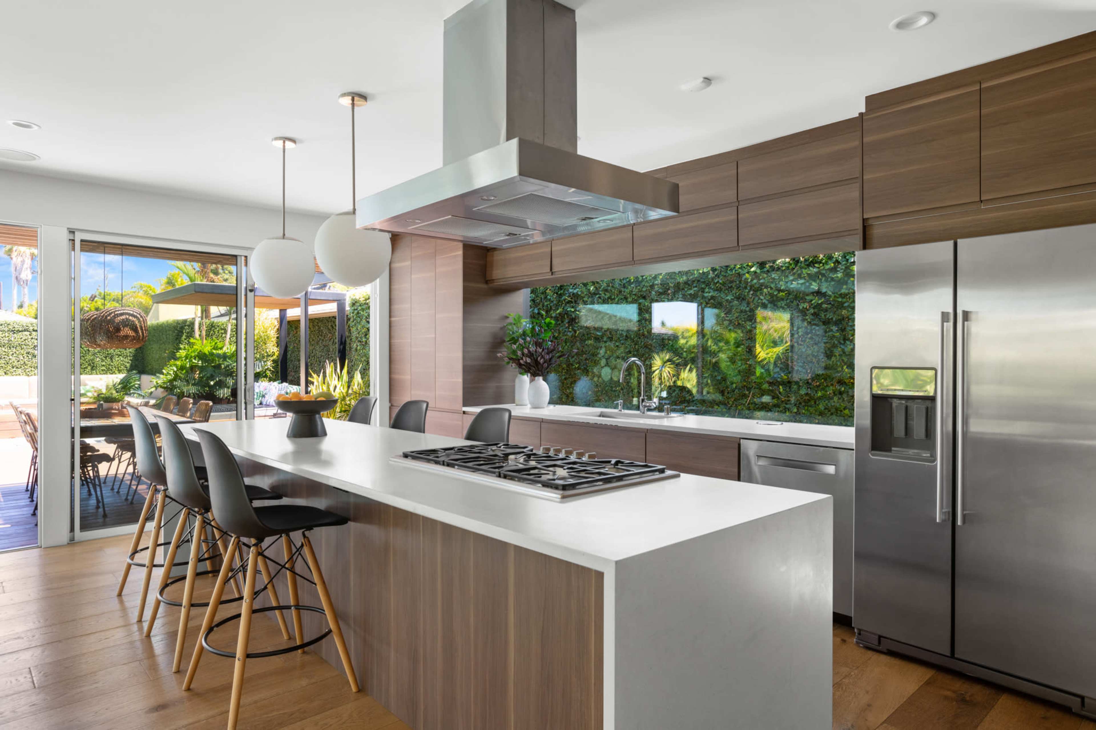 A modern kitchen with a large island, a stainless steel refrigerator, and a view of greenery through a window.