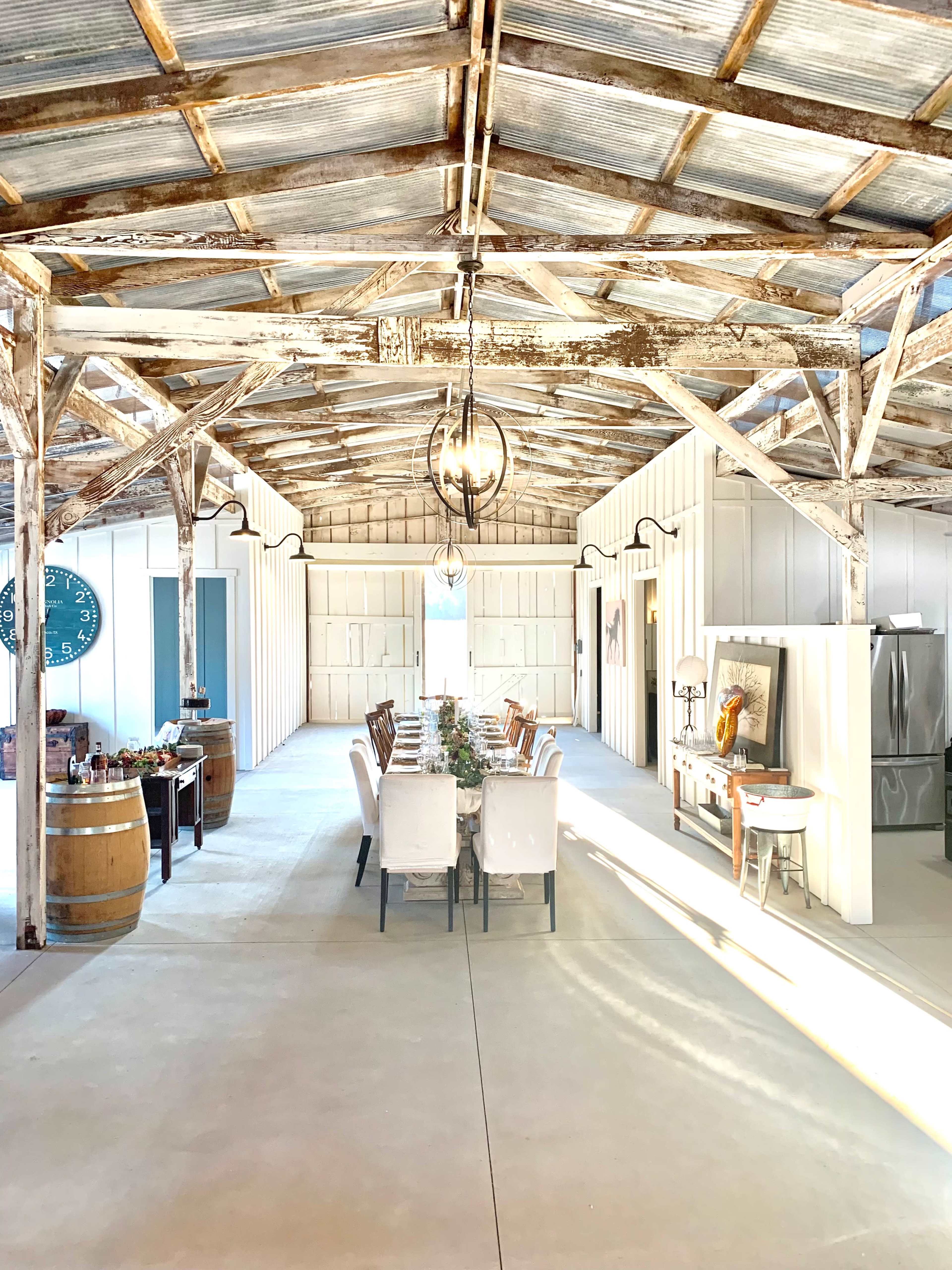 A spacious, rustic dining area with a long table set for a meal, surrounded by wooden barrels and bright natural light filtering through the open space.