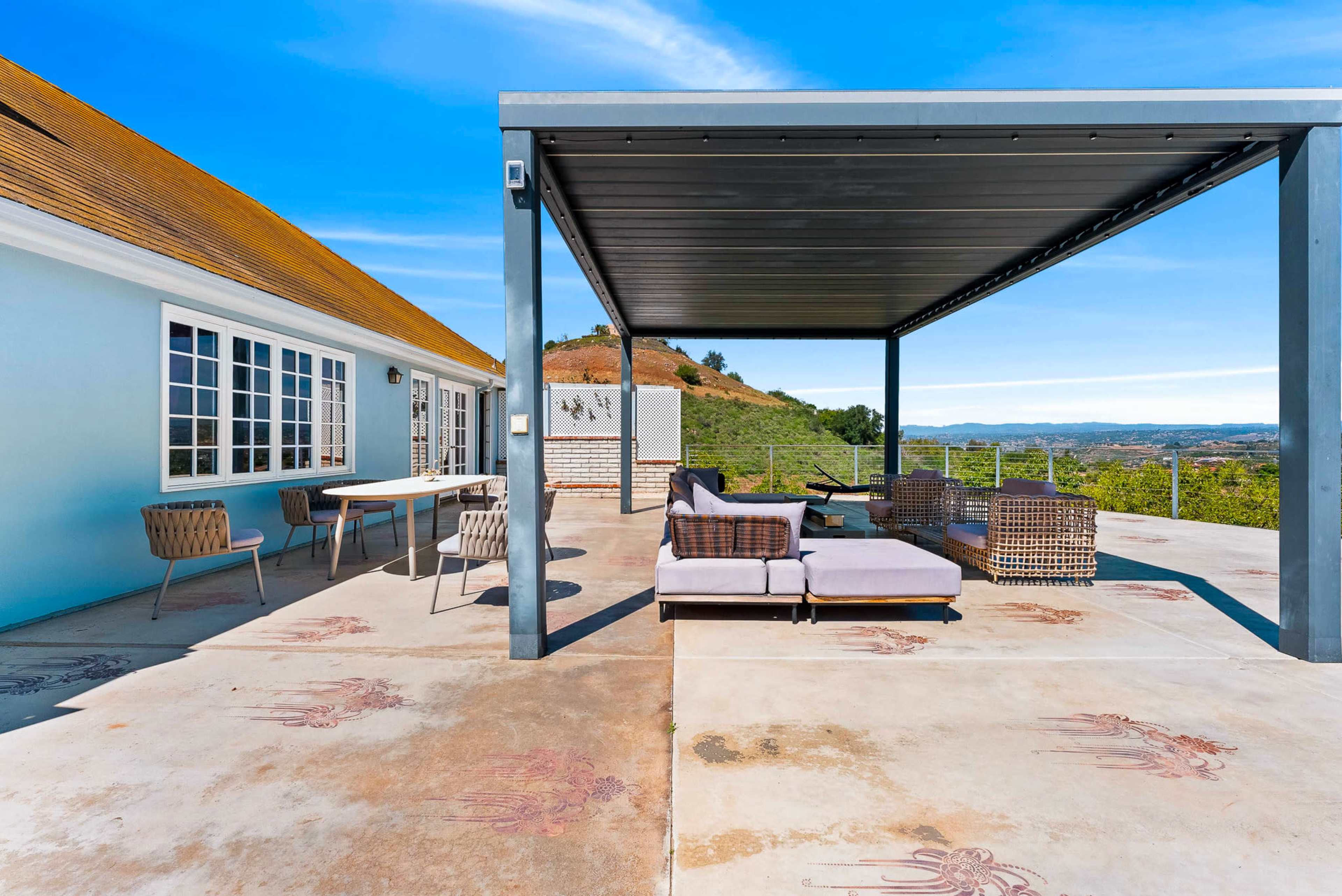 The image shows a patio area with outdoor seating under a shaded structure, overlooking a scenic landscape.