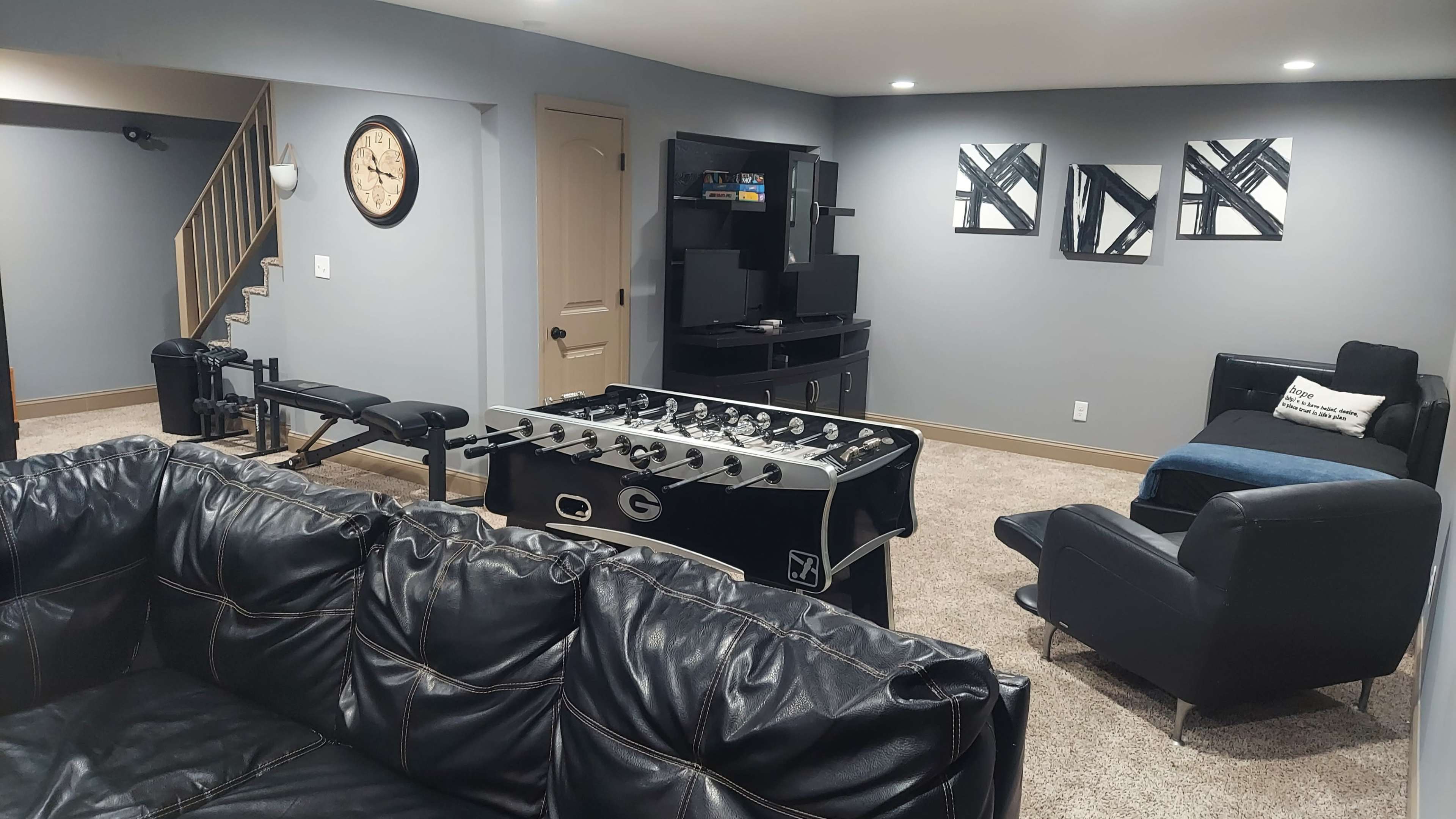 A modern basement recreation room featuring a foosball table, a television setup, a workout bench, and seating areas with black leather furniture.