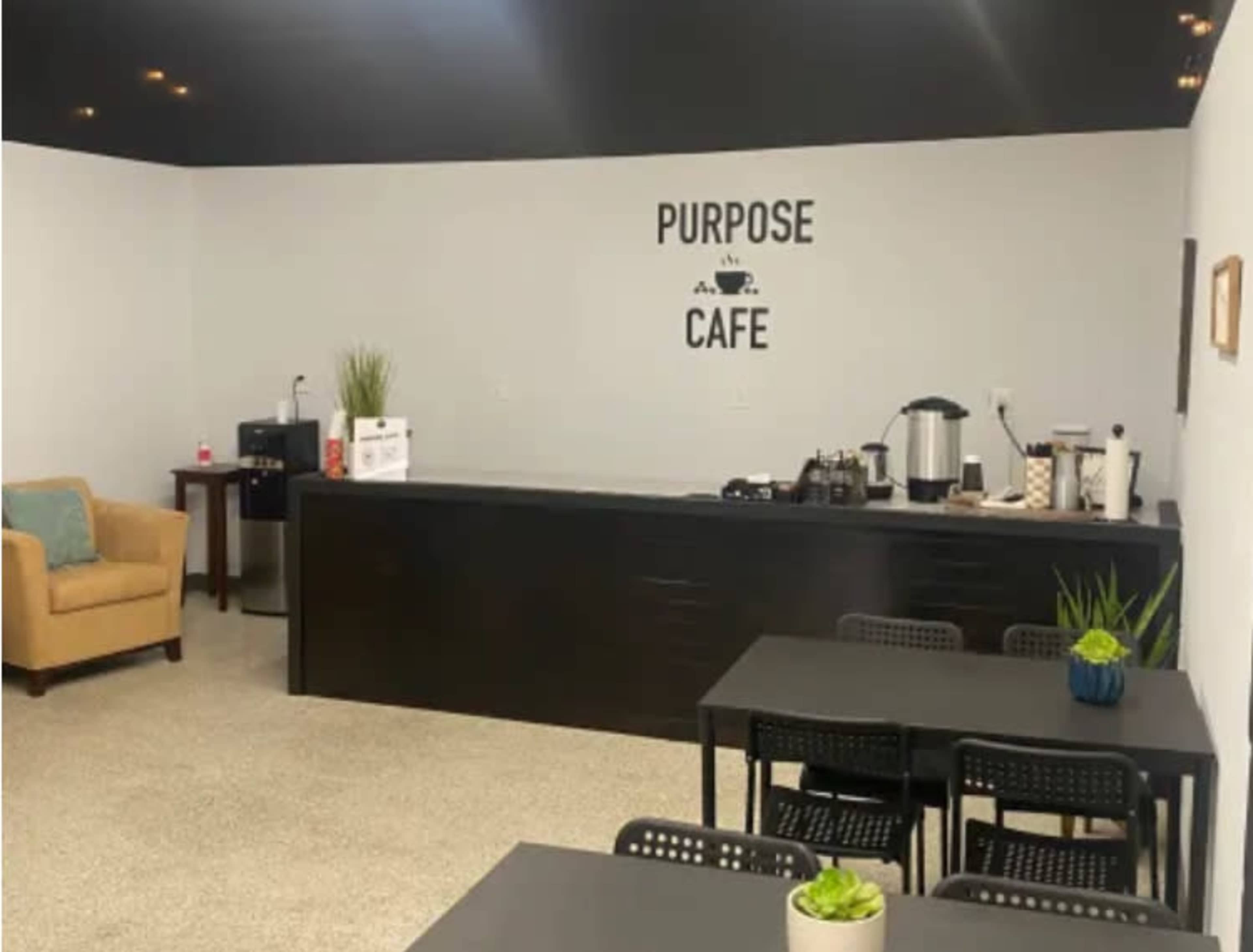 A simple cafe interior featuring a countertop with coffee equipment, tables with potted plants, and the words "PURPOSE CAFE" on the wall.
