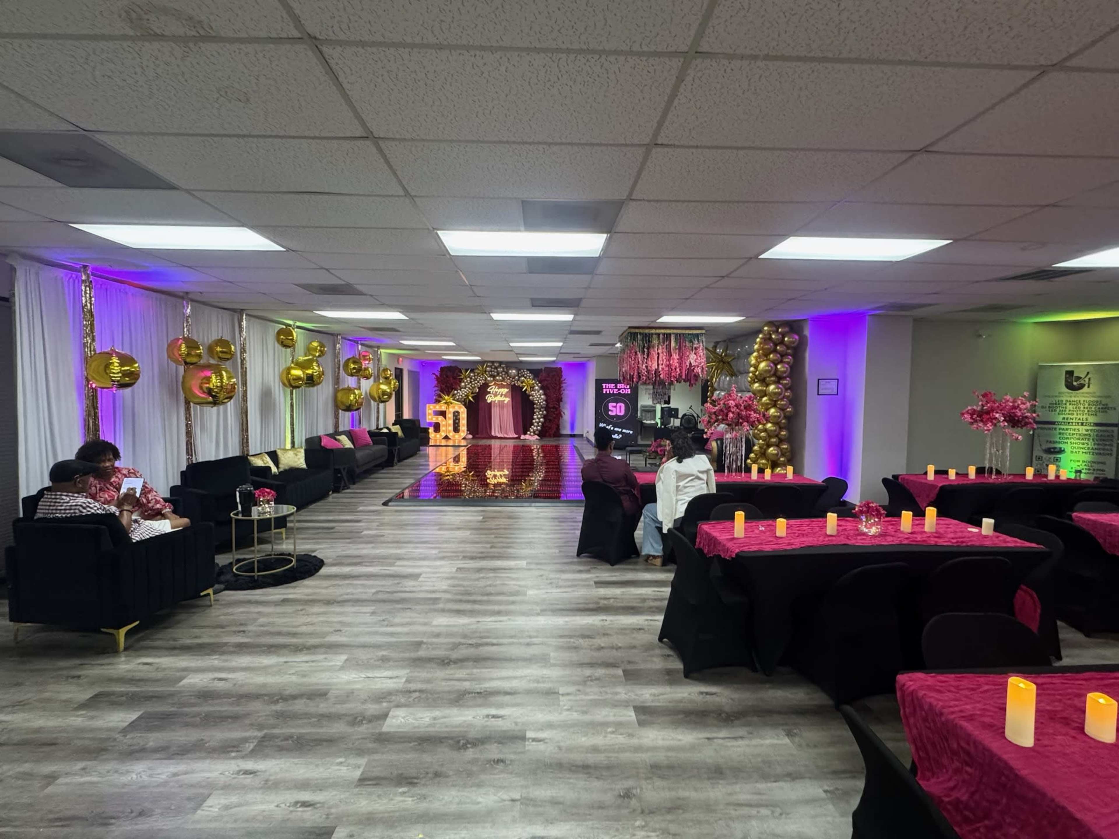 A decorated indoor event space features tables with dark tablecloths and pink centerpieces, along with gold balloons and a mirrored backdrop.