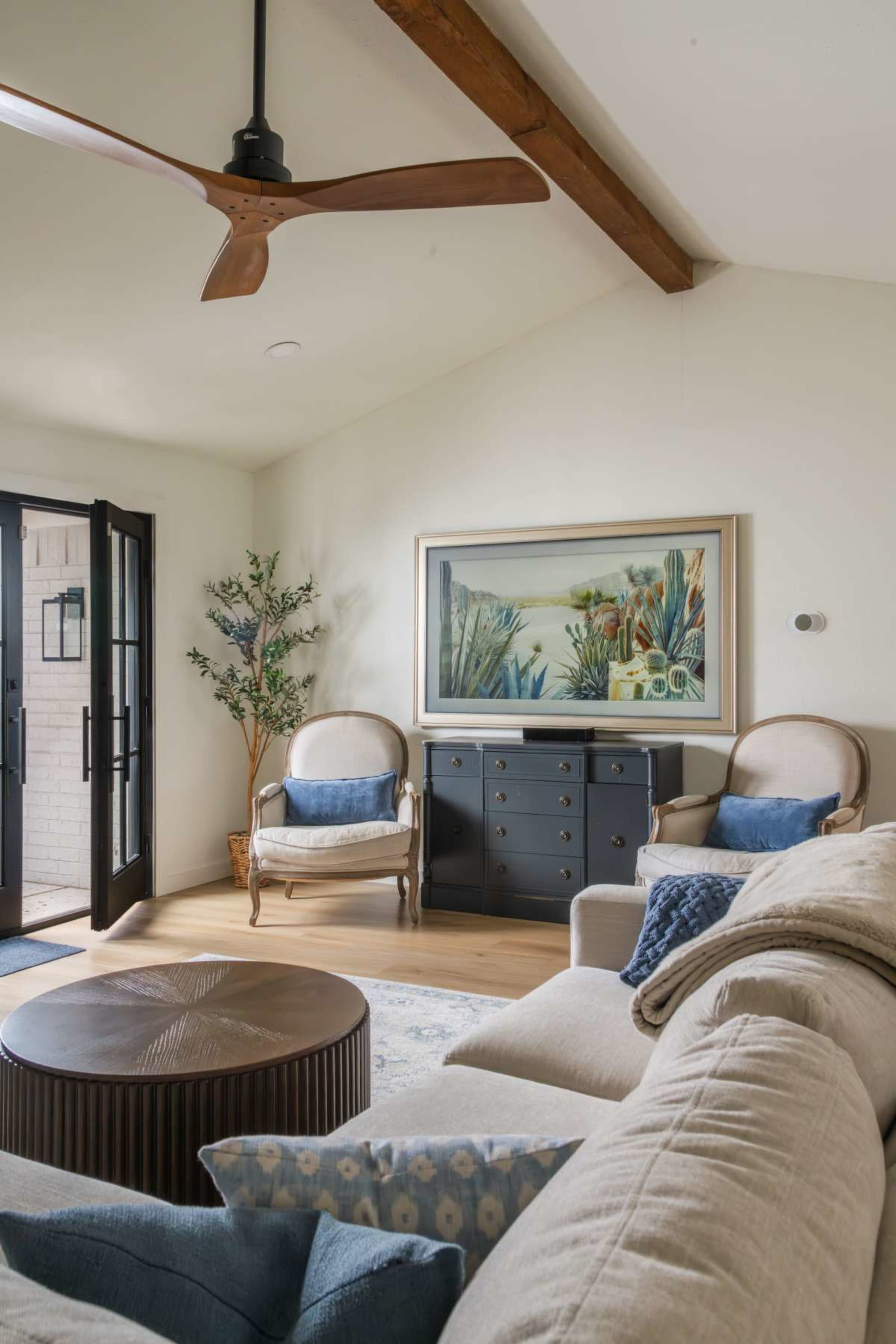 The image showcases a living room with a ceiling fan, two armchairs, a dark chest of drawers, and a large framed painting.