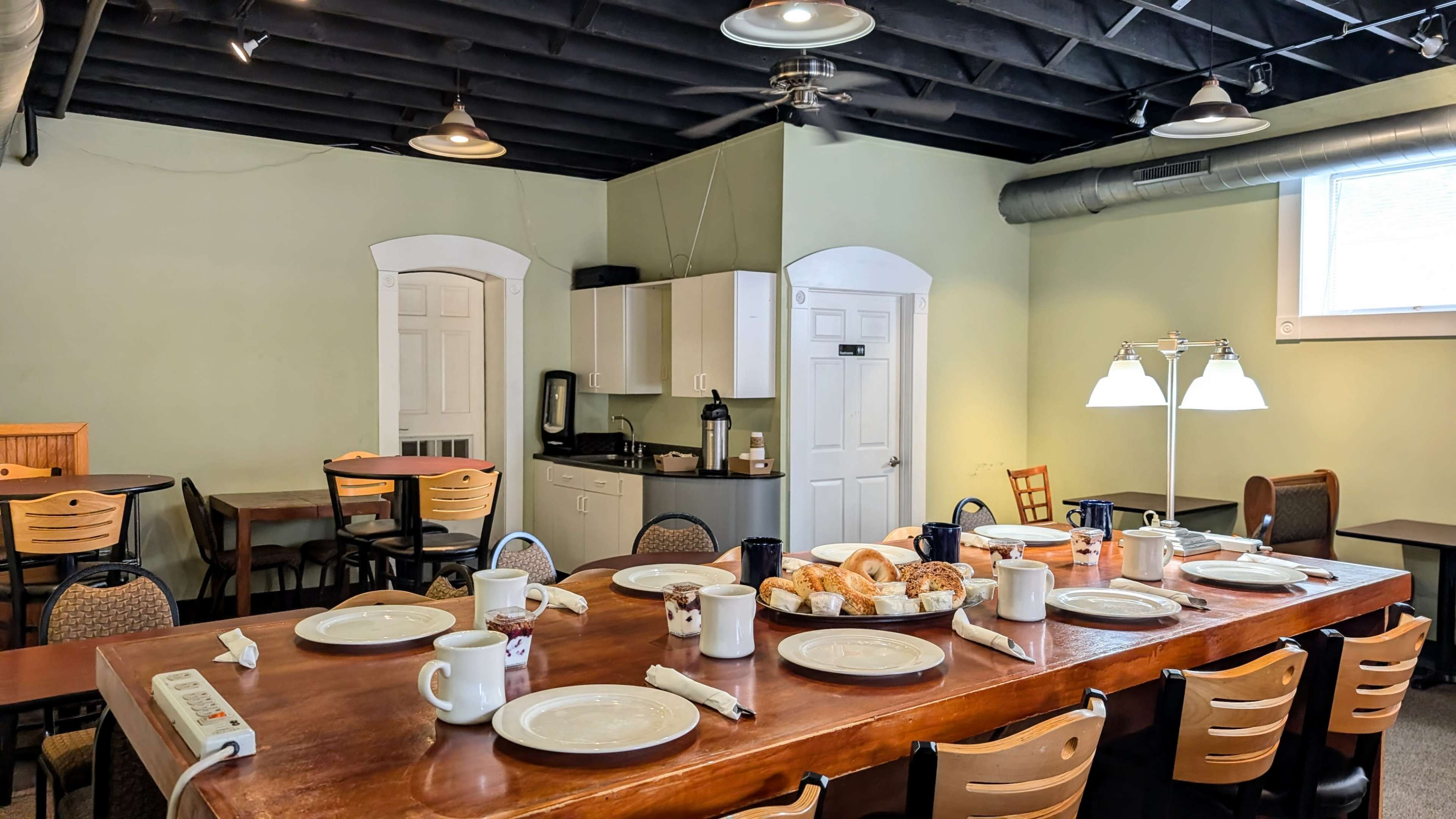 The image shows a dining area with a large wooden table set for a meal, surrounded by chairs, and a kitchenette in the background.