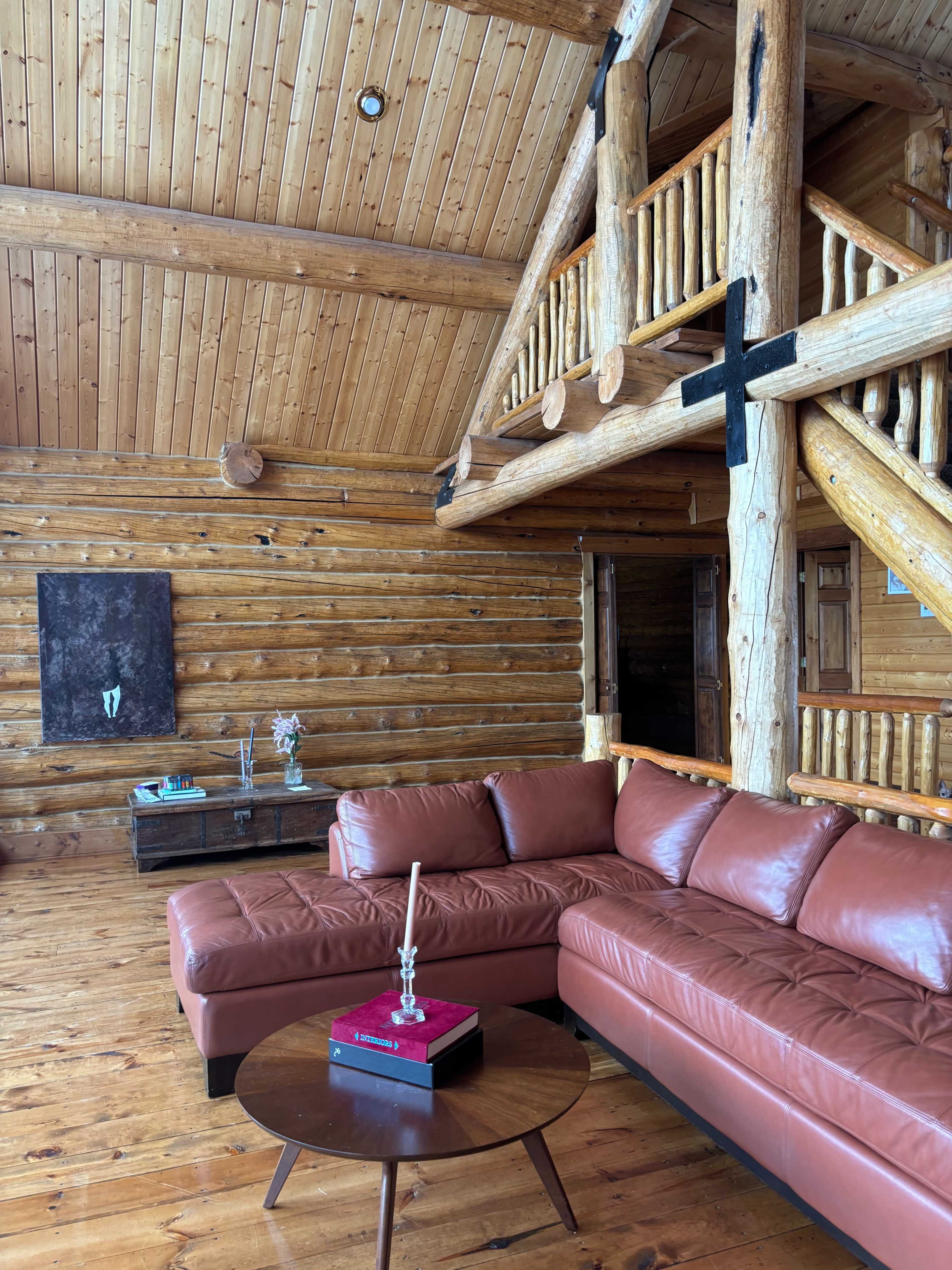 The interior of a log cabin features a brown leather sectional sofa, a round coffee table with a decorative item on it, and wooden beams along the ceiling.