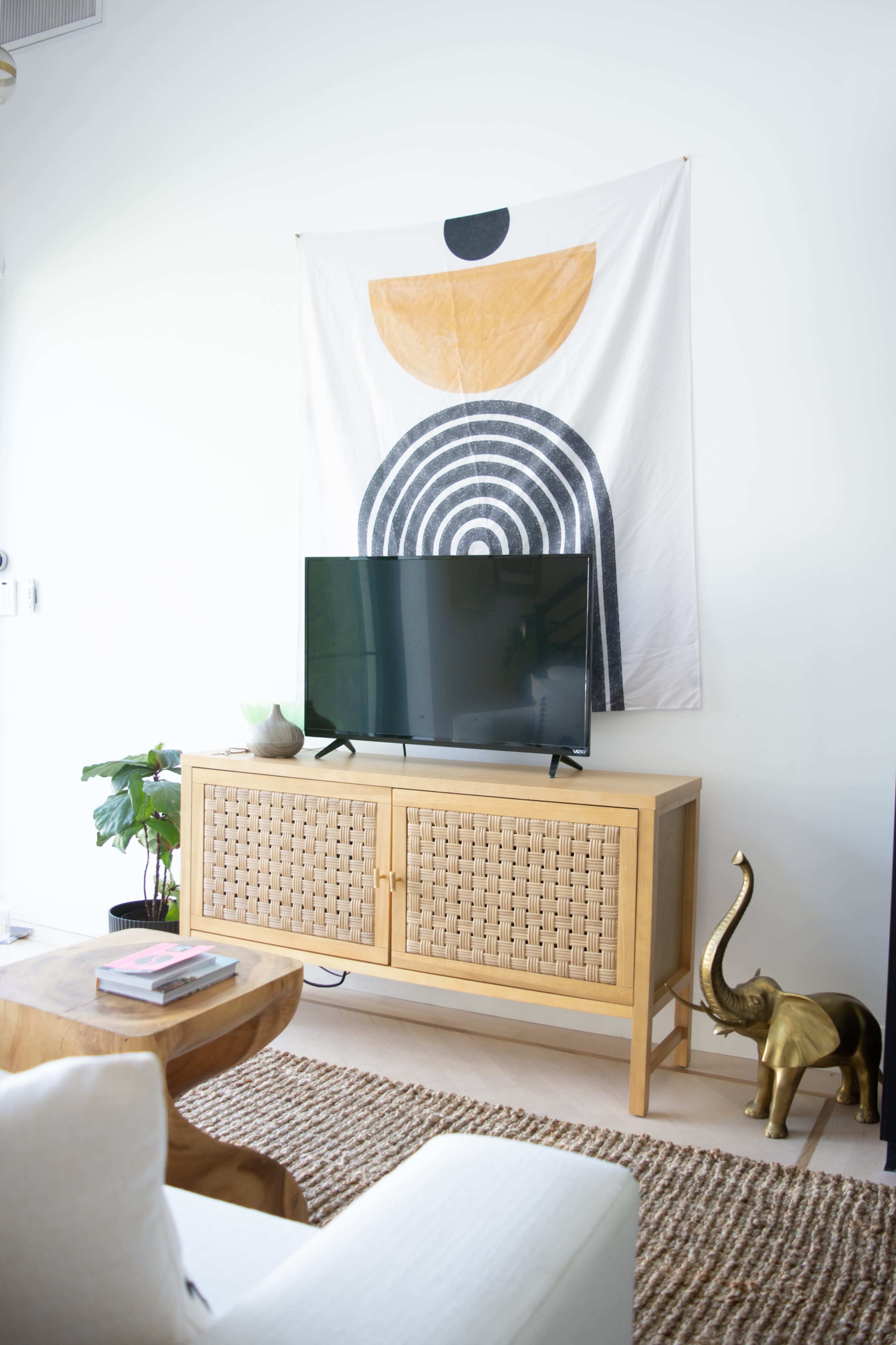 A living room with a wooden cabinet holding a television, a geometric wall hanging, a plant, and a decorative elephant figurine.
