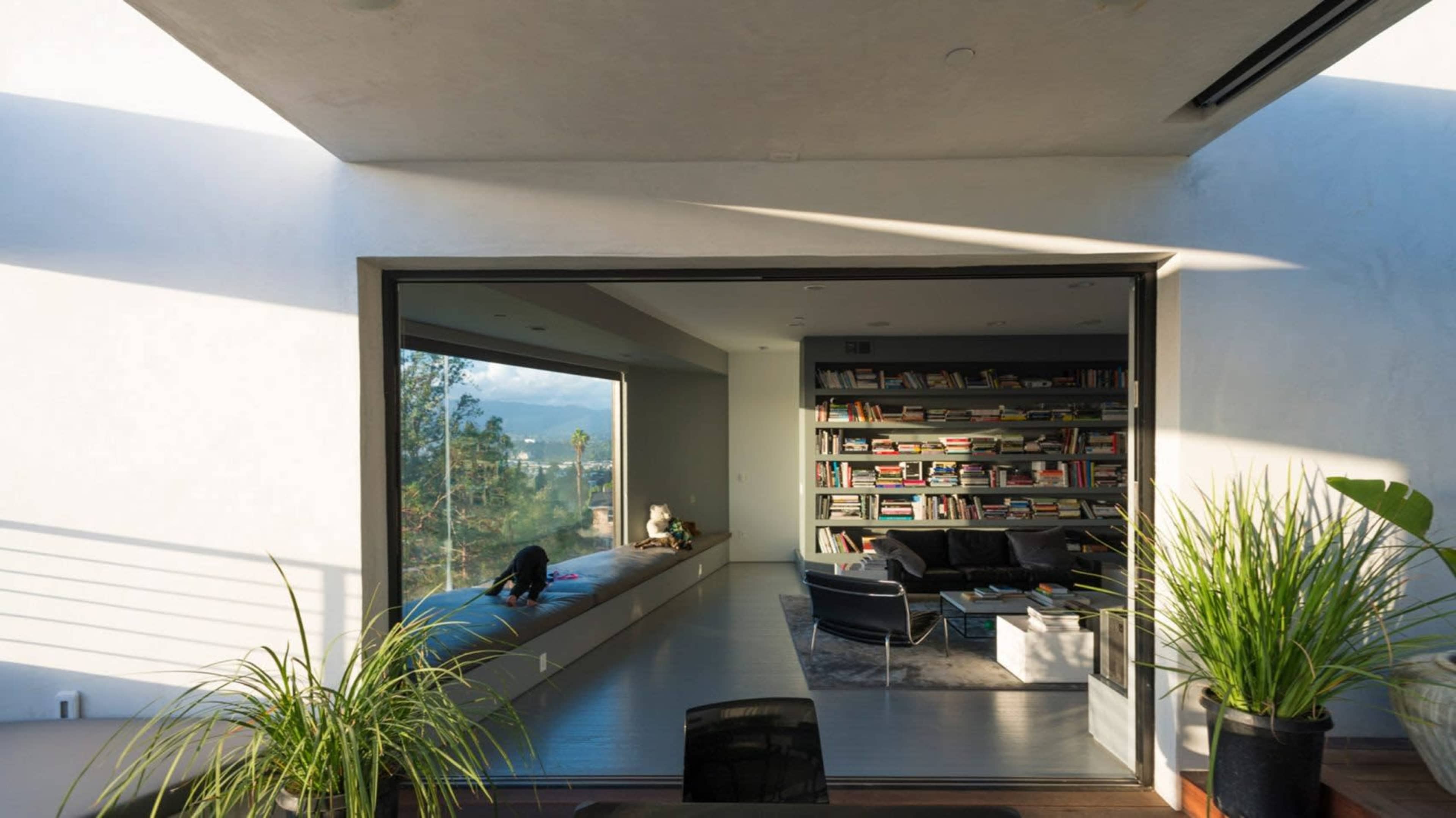 A spacious interior features a large window with a view of mountains, a bookshelf filled with books, and plants in the foreground.