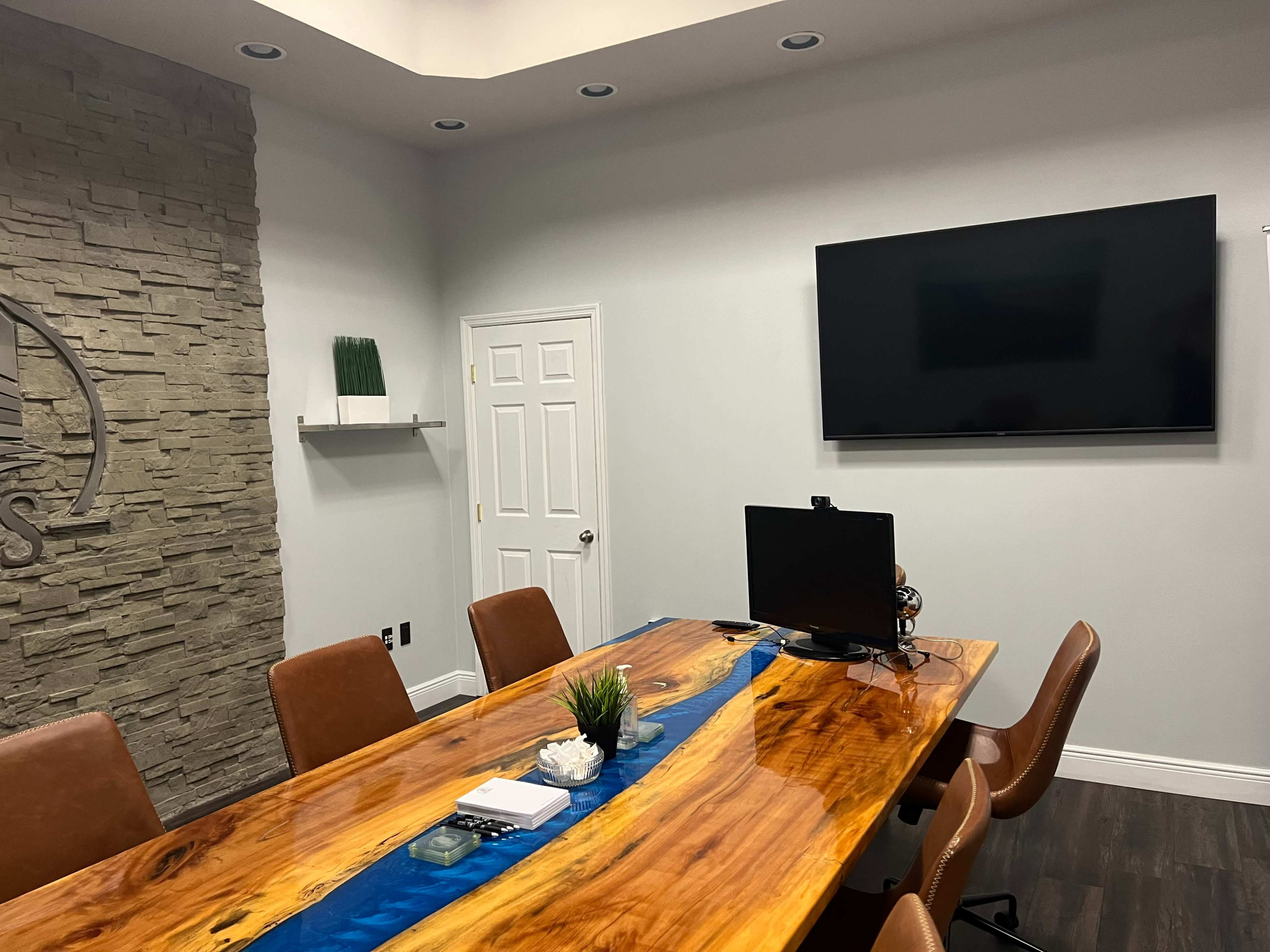 A modern conference room features a long wooden table with a blue resin inlay, a television mounted on the wall, and a minimalist decor.