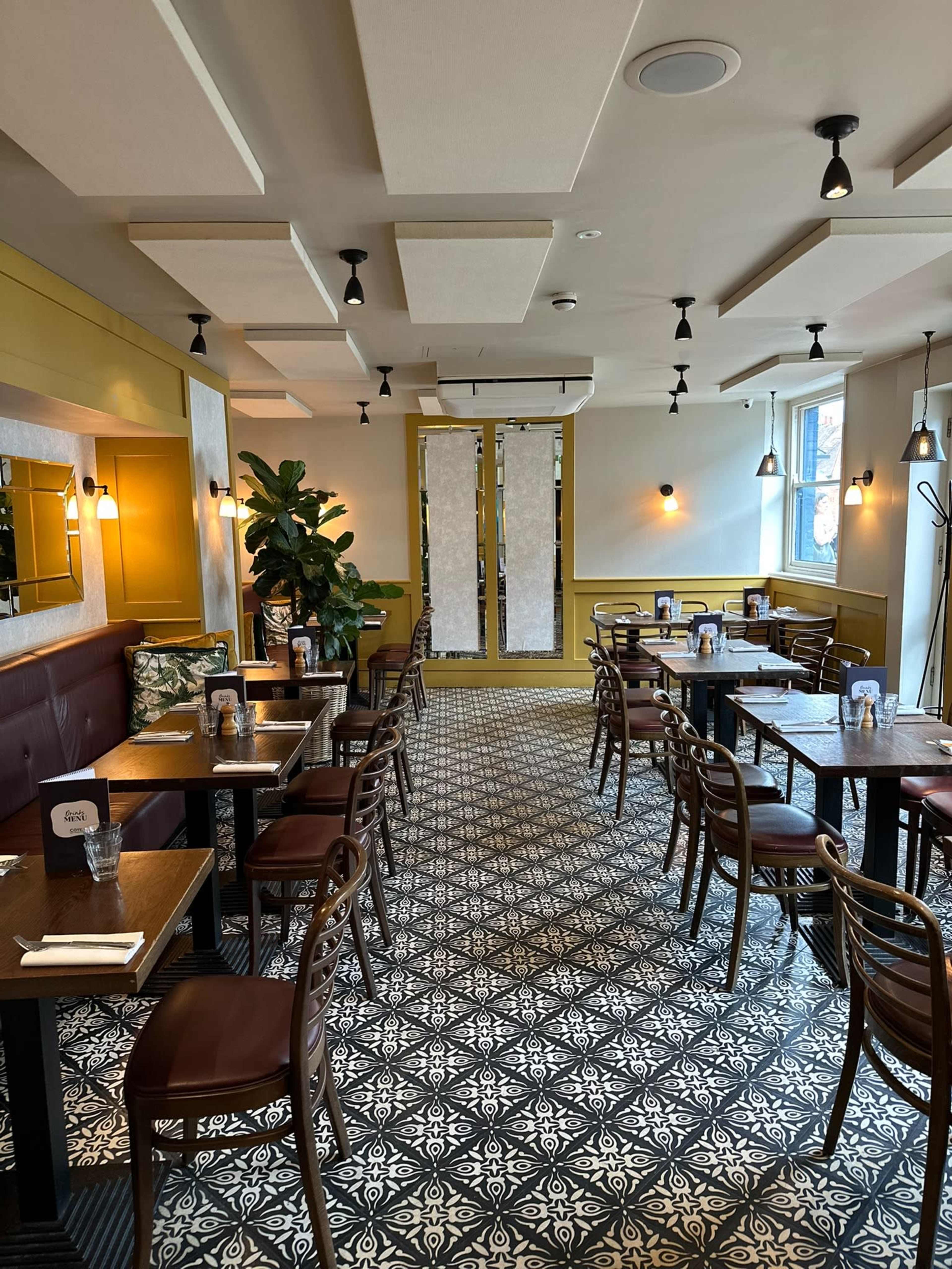 The image shows a spacious restaurant interior with patterned tile flooring, wooden tables, and chairs arranged around the room, featuring yellow walls and greenery.