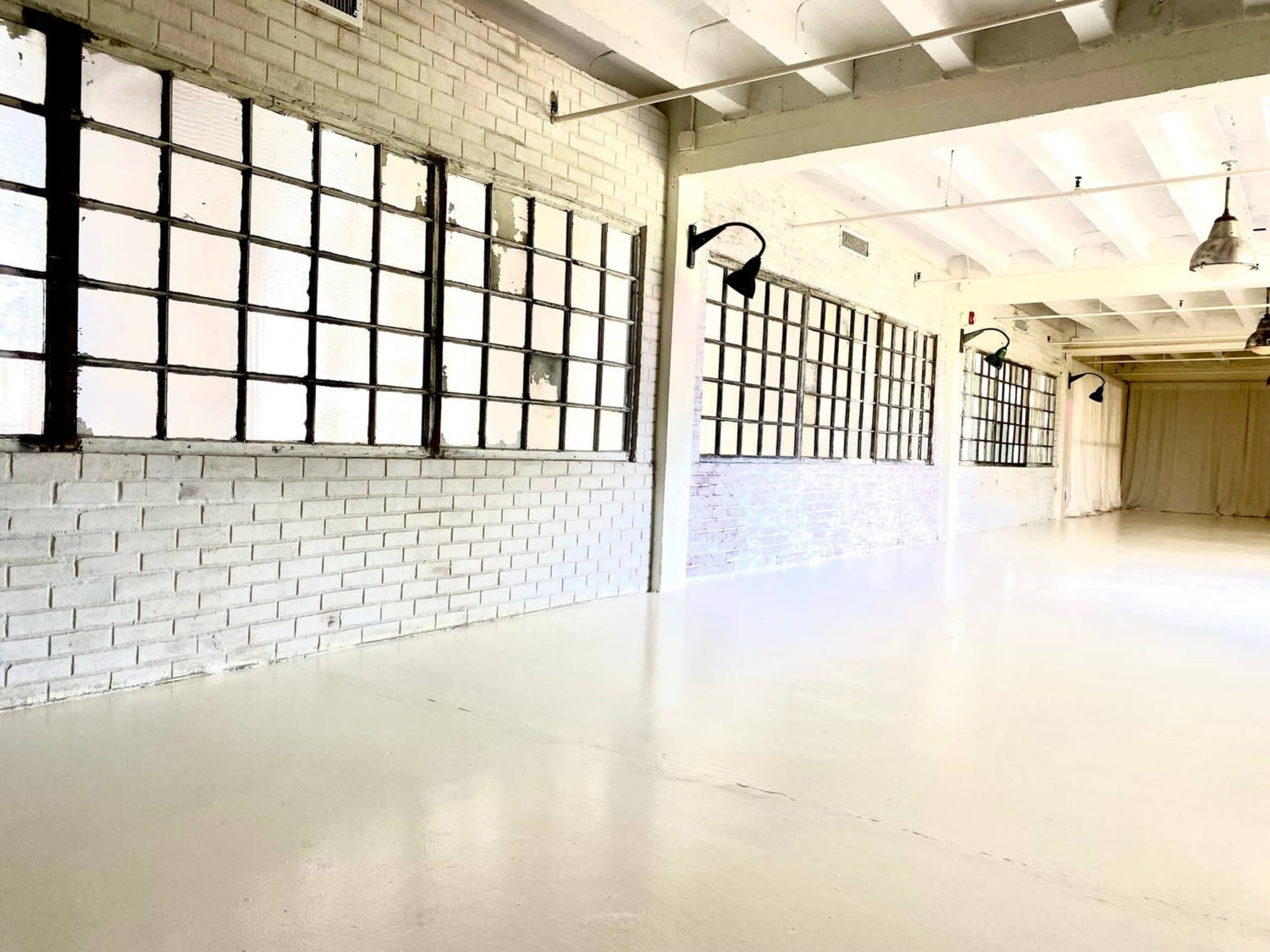 An empty, well-lit interior space with white brick walls and large, divided windows along one side.