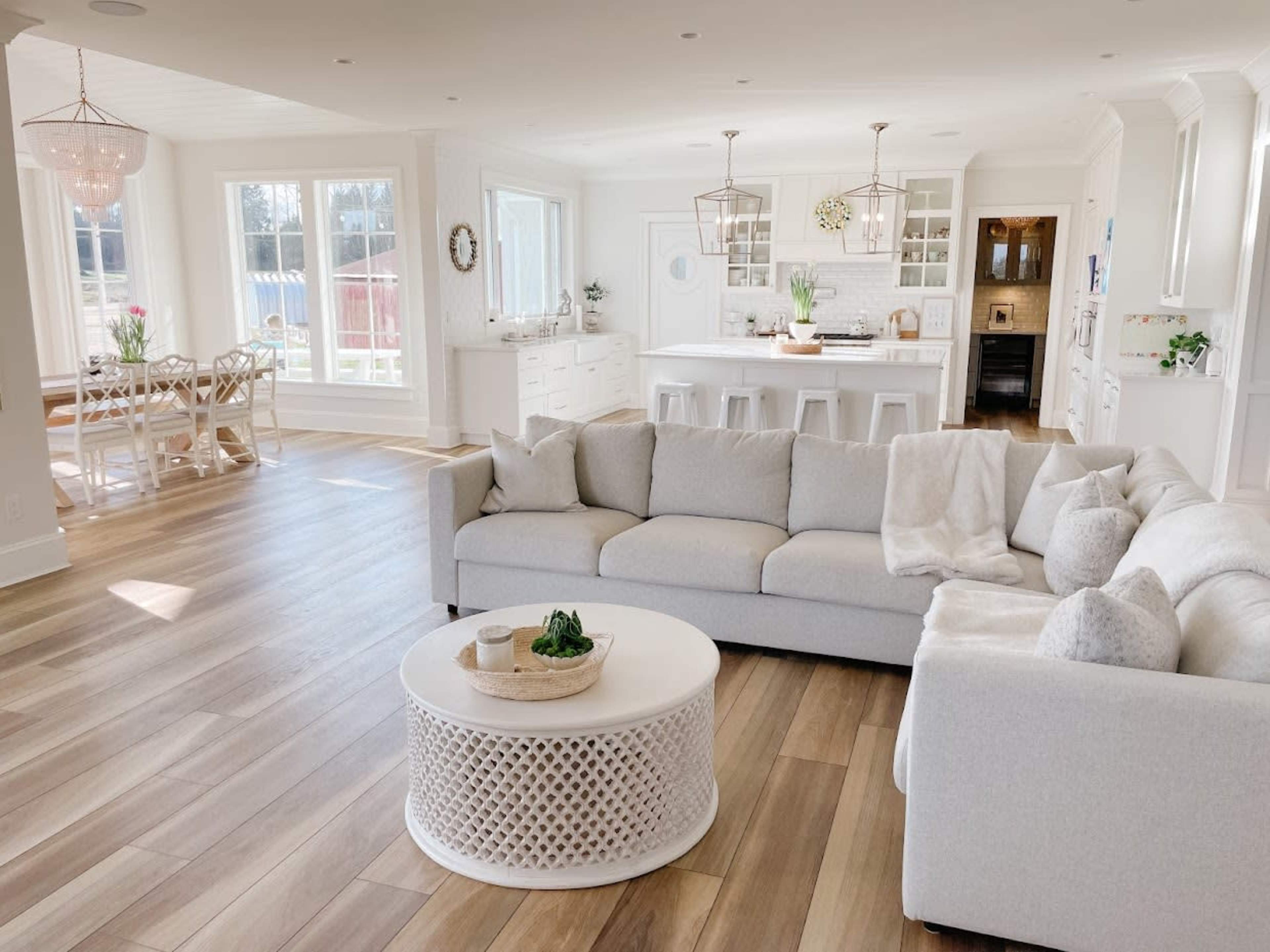 A spacious living area features a light-colored sectional sofa, a round coffee table, and large windows allowing natural light to fill the room.