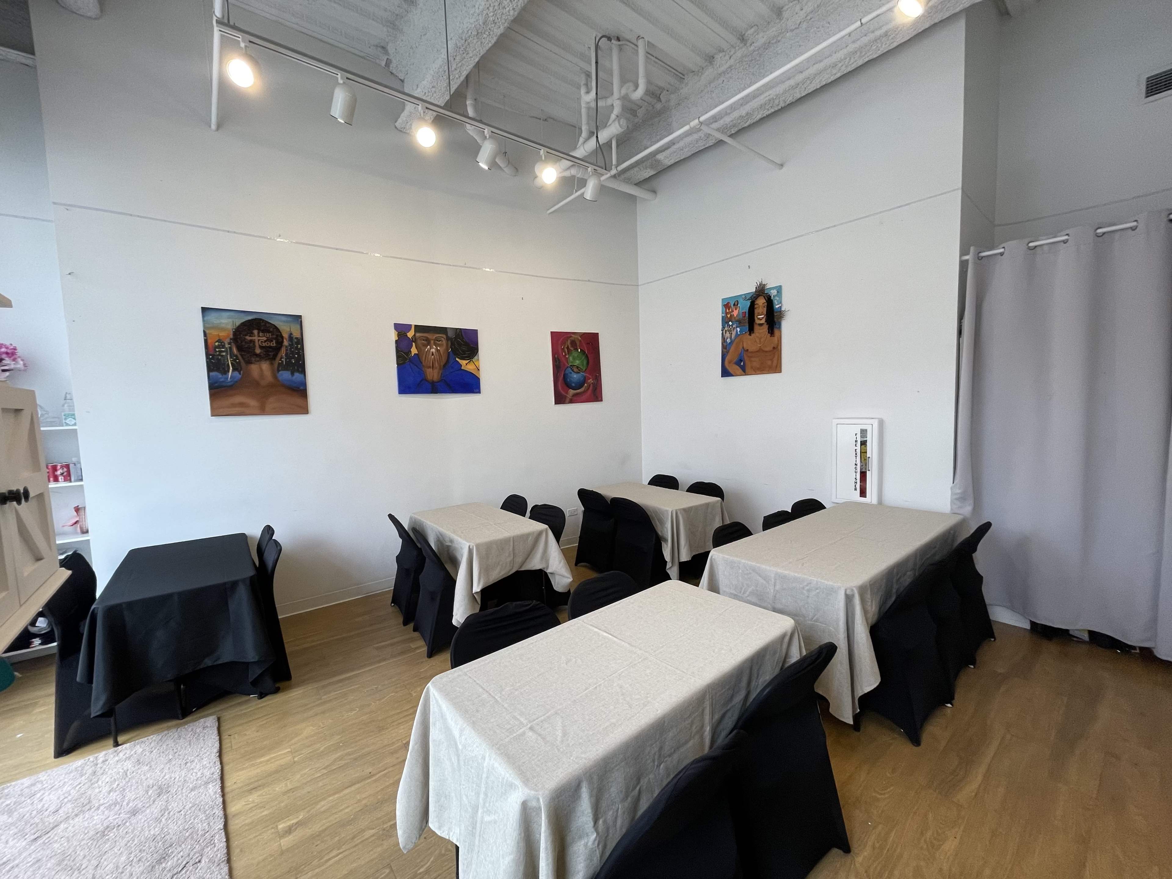 The image shows a brightly lit room set up with several tables covered in light-colored tablecloths and adorned with black chairs, featuring colorful paintings on the walls.