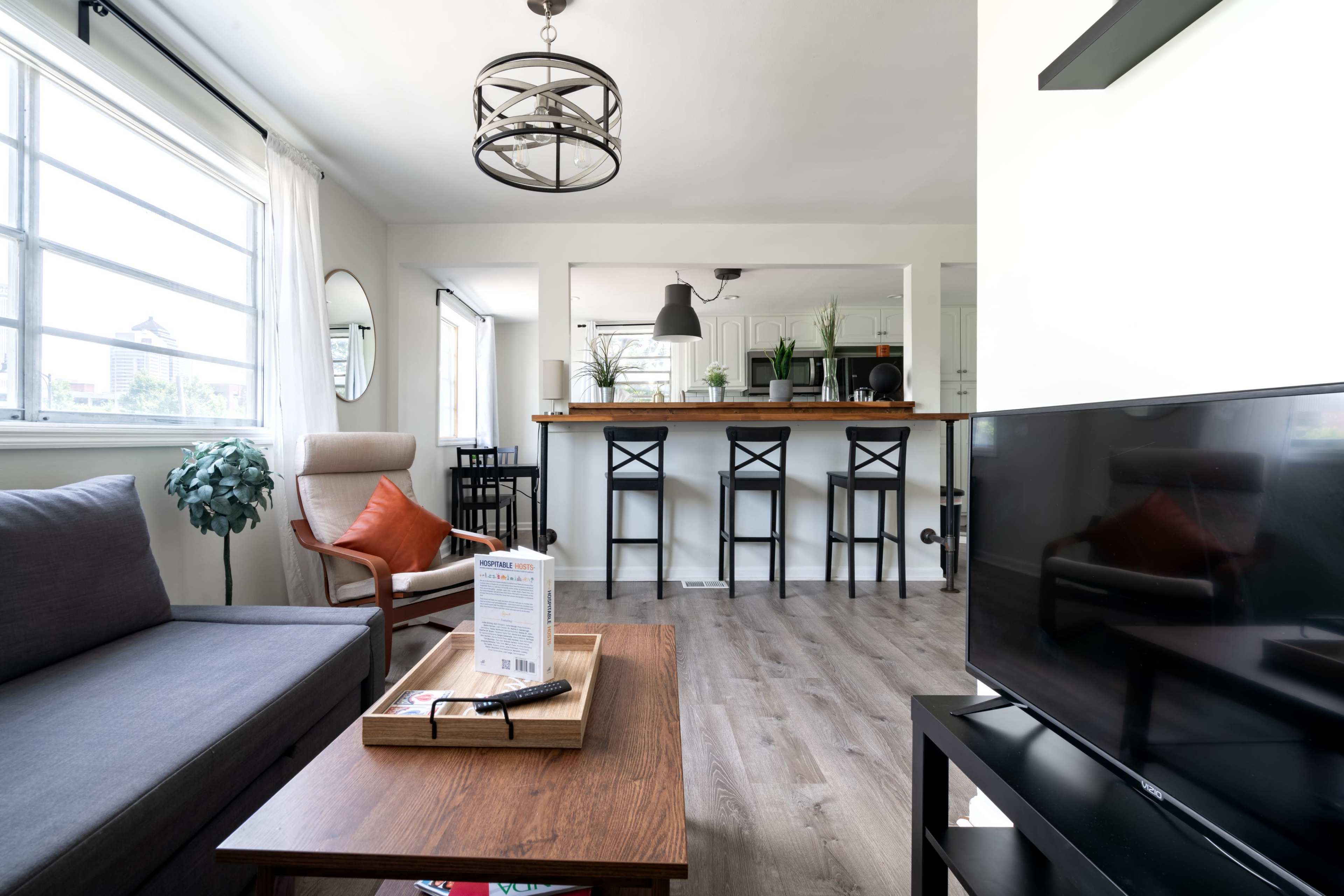A cozy living room features a gray sofa, a wooden coffee table with a remote, and a television, with an open kitchen area in the background.