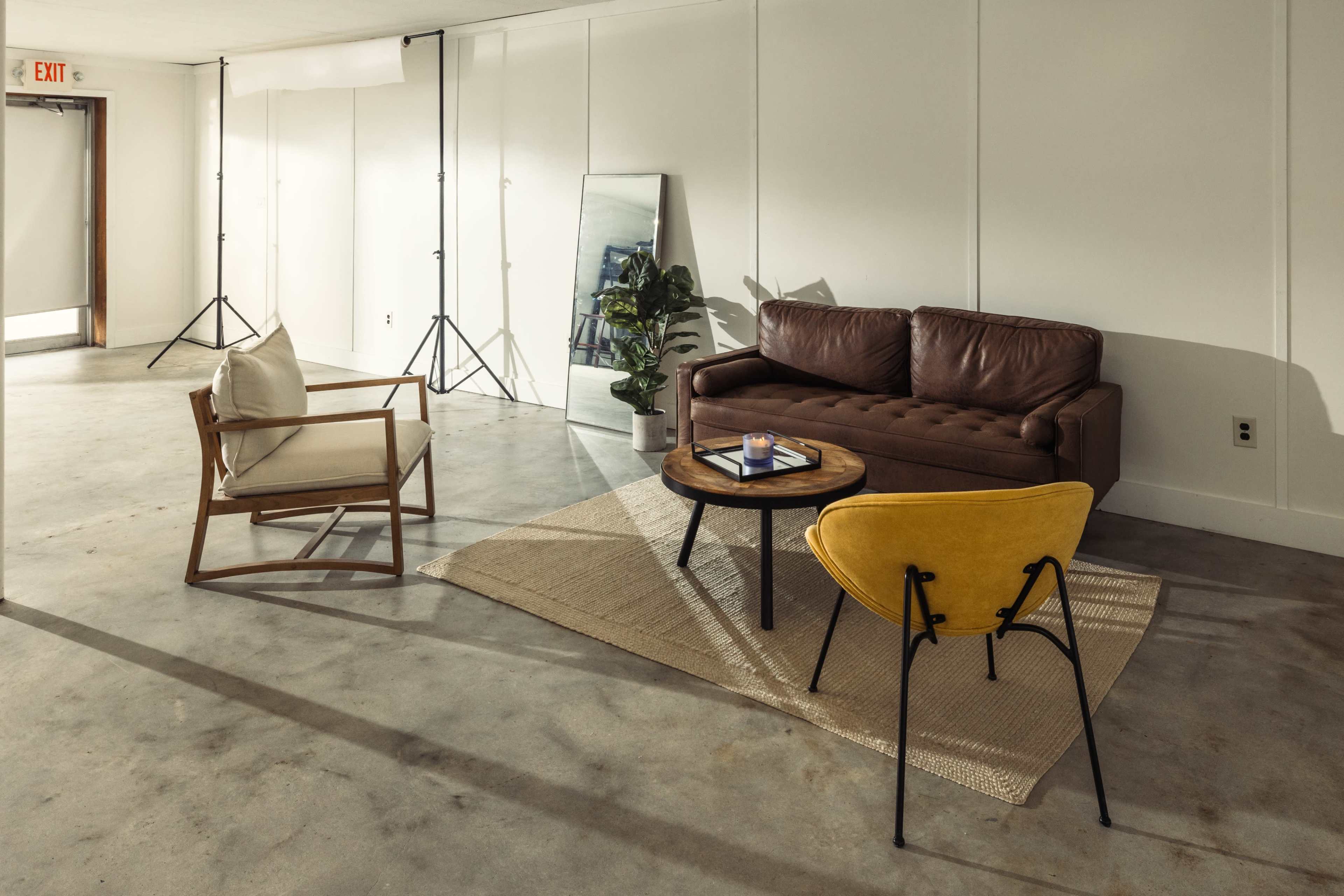 The image shows a minimalist living space with a brown leather sofa, a wooden chair, a round coffee table, and a yellow accent chair on a textured rug.
