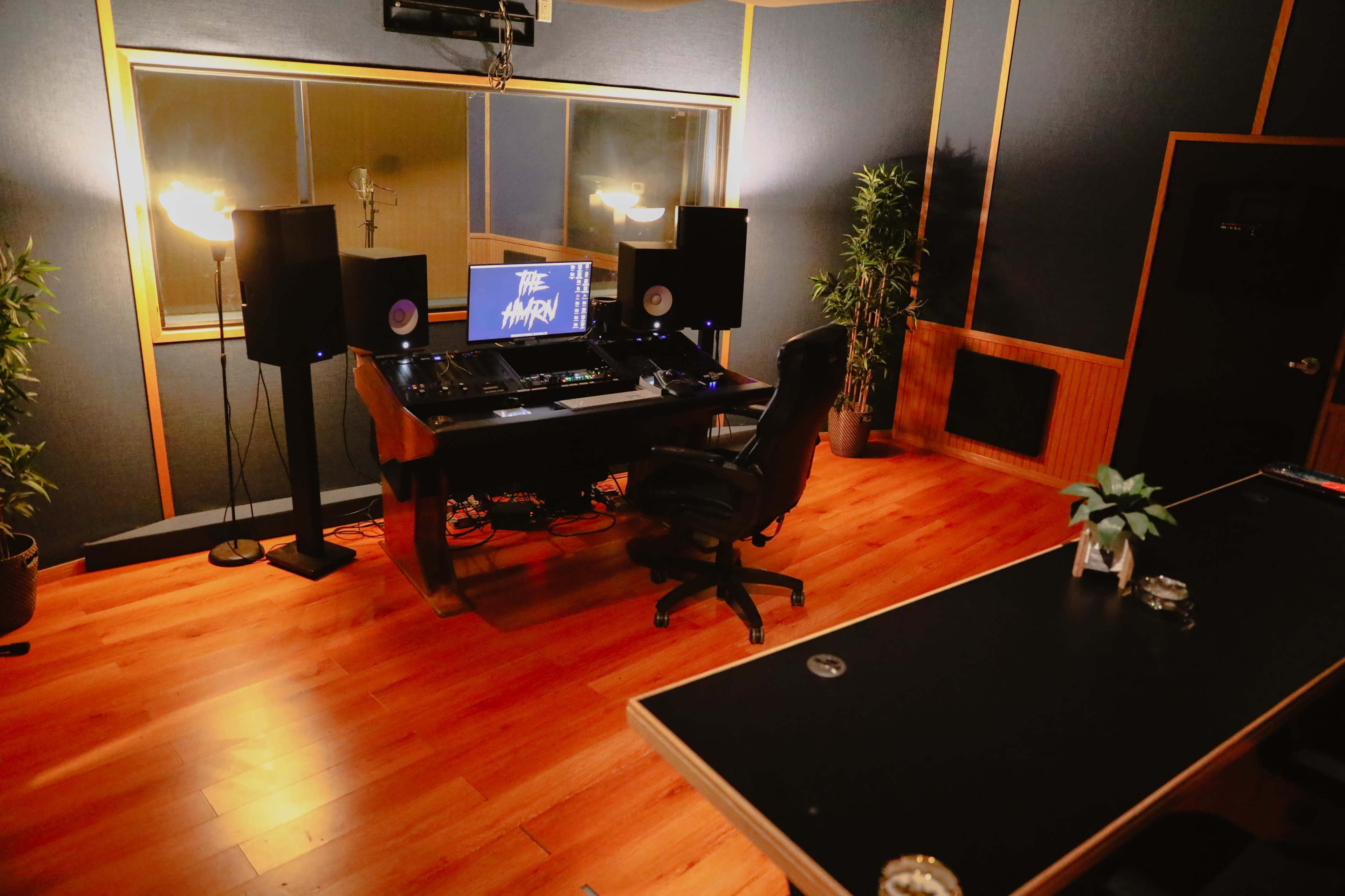 The image shows a modern music studio with a central workstation, large speakers, and a long table surrounded by plants, all set against blue walls and wooden flooring.